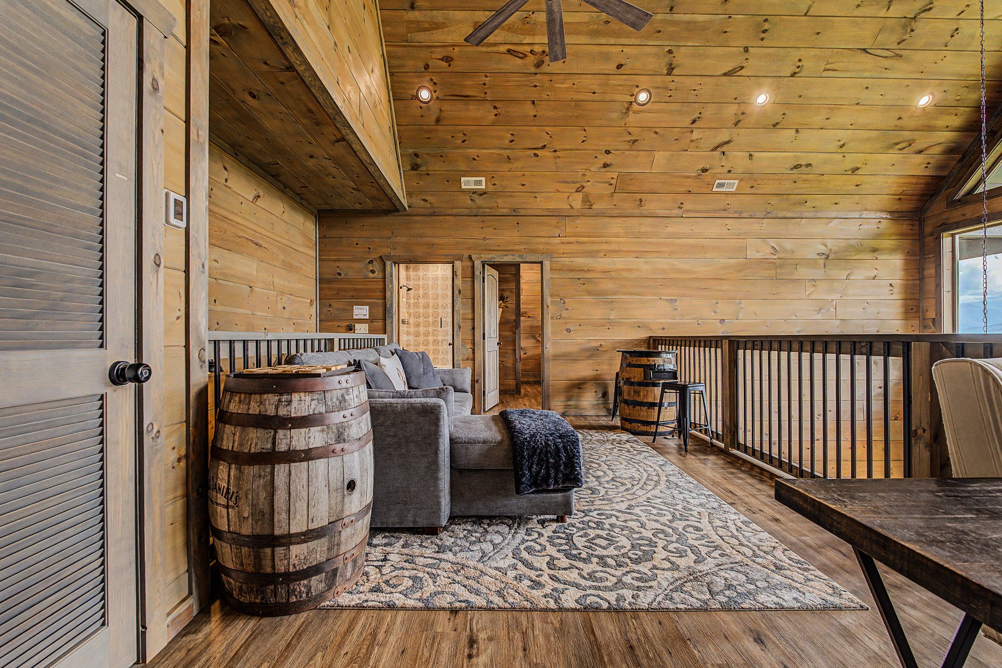 A cozy interior room with wood-paneled walls and ceiling. There is a gray couch with pillows, a patterned rug, a wooden barrel table, and a black stool. A ceiling fan and recessed lighting are visible, and there is a window on one side.