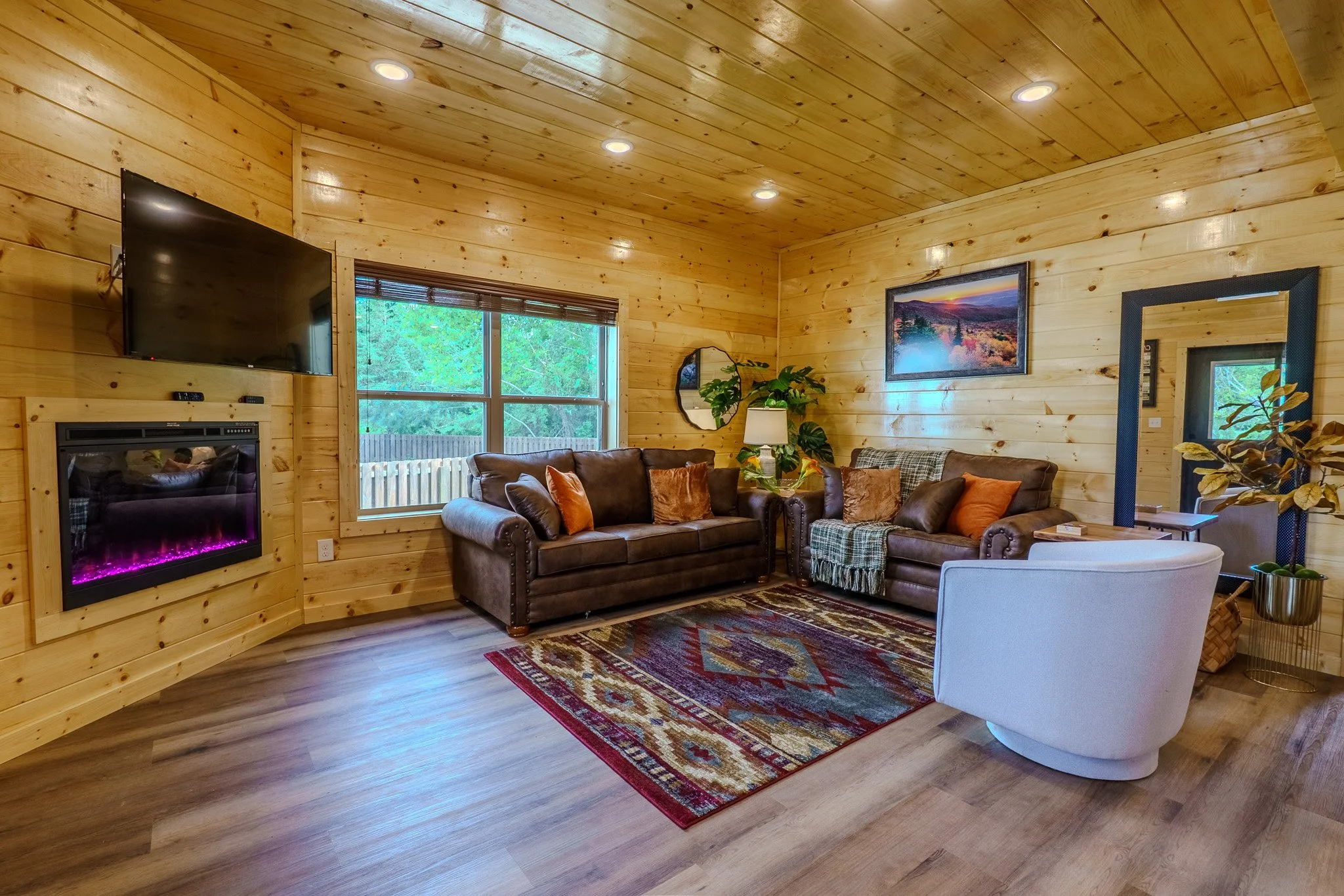 Living room with wood-paneled walls, hardwood floor, two brown sofas with pillows, a white armchair, a colorful area rug, a large window with greenery outside, a mounted TV, an electric fireplace, a lamp, wall art, and various plants.