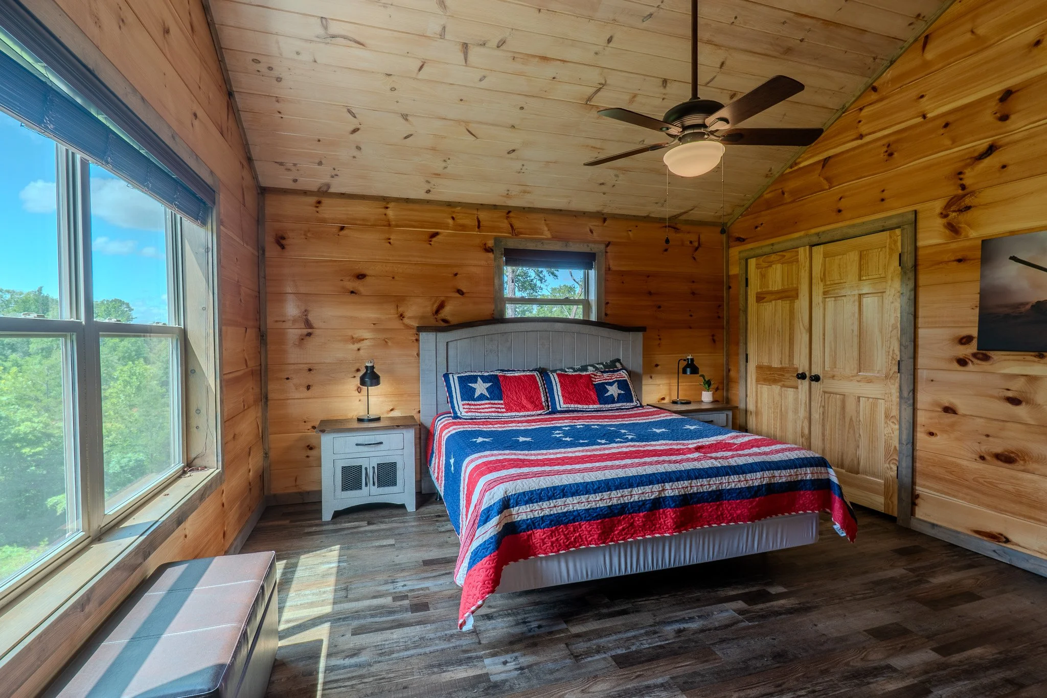 A bedroom with wooden panel walls and ceiling, featuring a bed with a red, white, and blue American flag-themed quilt and pillows, two nightstands with lamps, a window, a ceiling fan, a closet with wooden doors, and a framed picture on the wall.