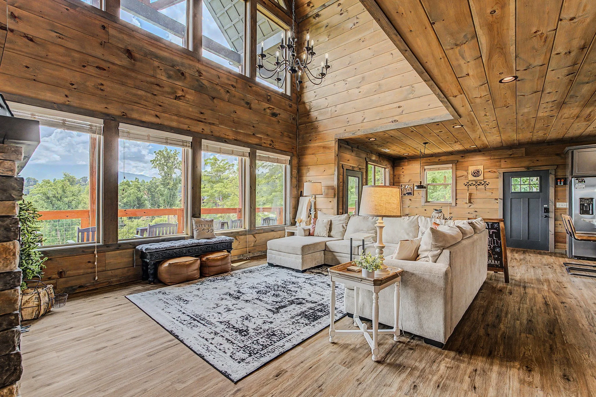 Living room with wood-paneled walls and ceiling, large windows showing a scenic mountain view, a white sectional sofa with throw pillows, a black and white patterned area rug, a wooden coffee table, lamps, and decorative wall art.