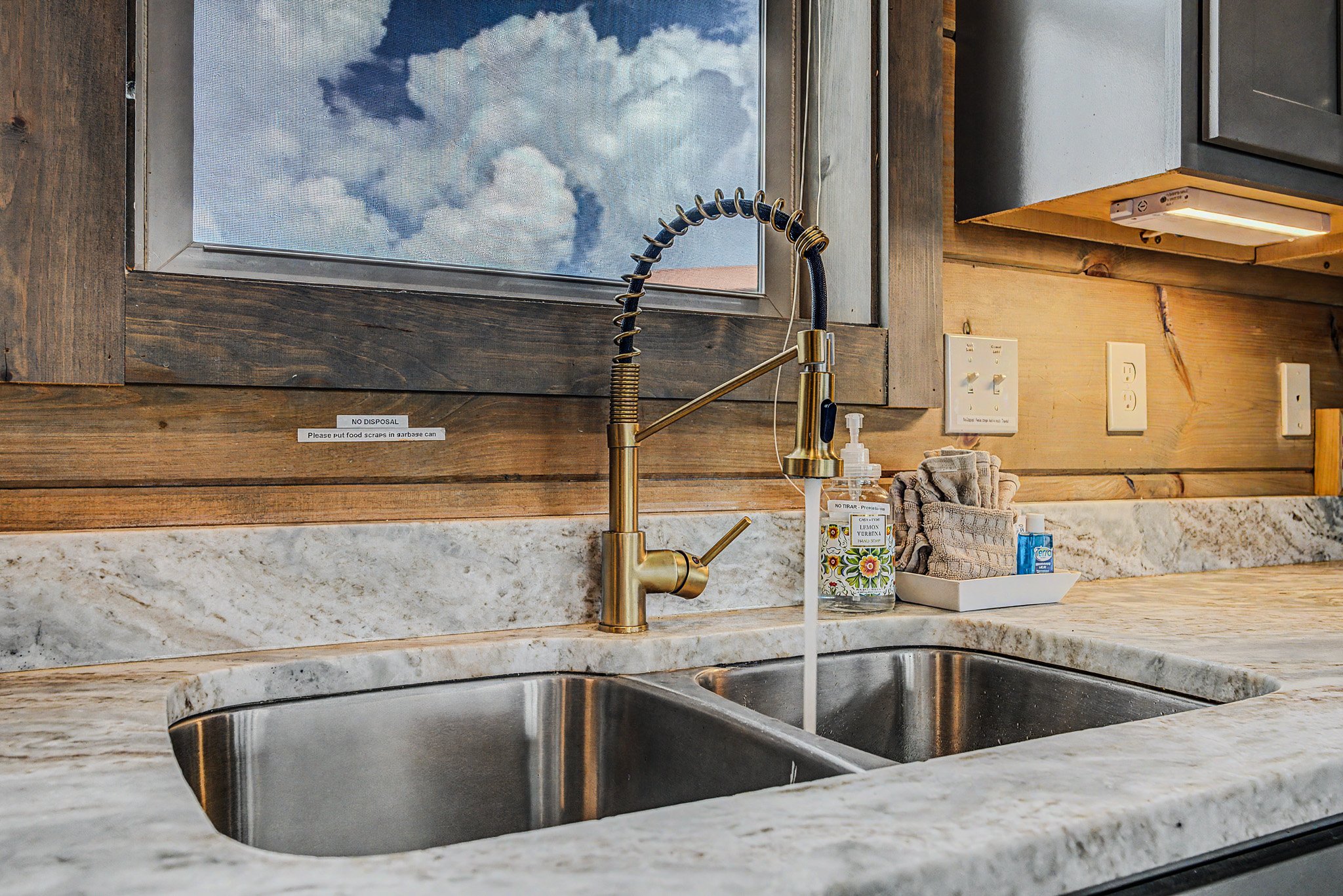Kitchen sink with a gold-colored faucet, set in a marble countertop beneath a window showing a cloudy sky. Next to the sink are containers, hand soap, a towel, and electrical outlets on a wooden wall.