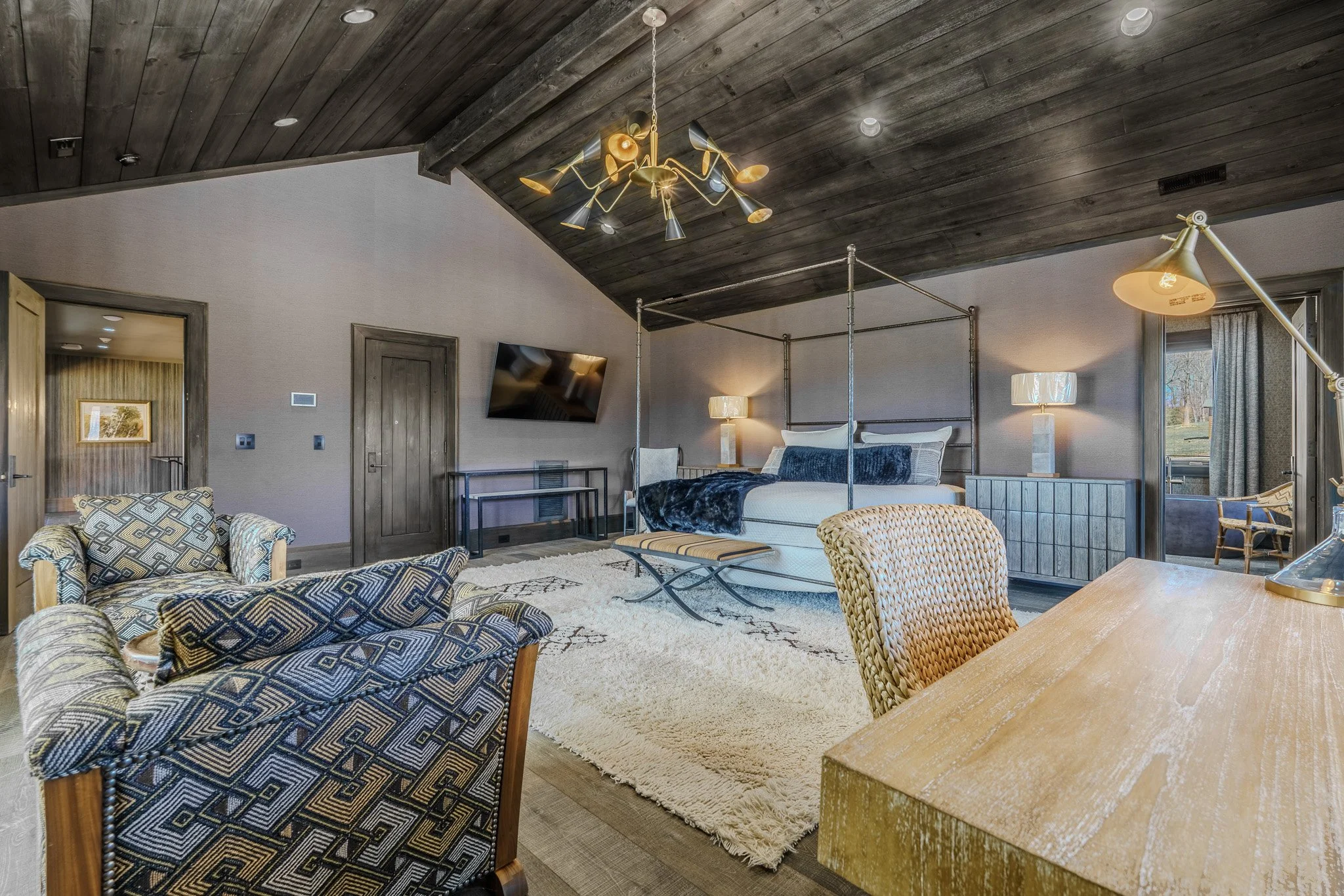 A spacious hotel room with a bed, armchairs, a desk, and a mounted flat-screen TV. The room has a wooden vaulted ceiling with modern light fixtures, and large windows with curtains letting in natural light.