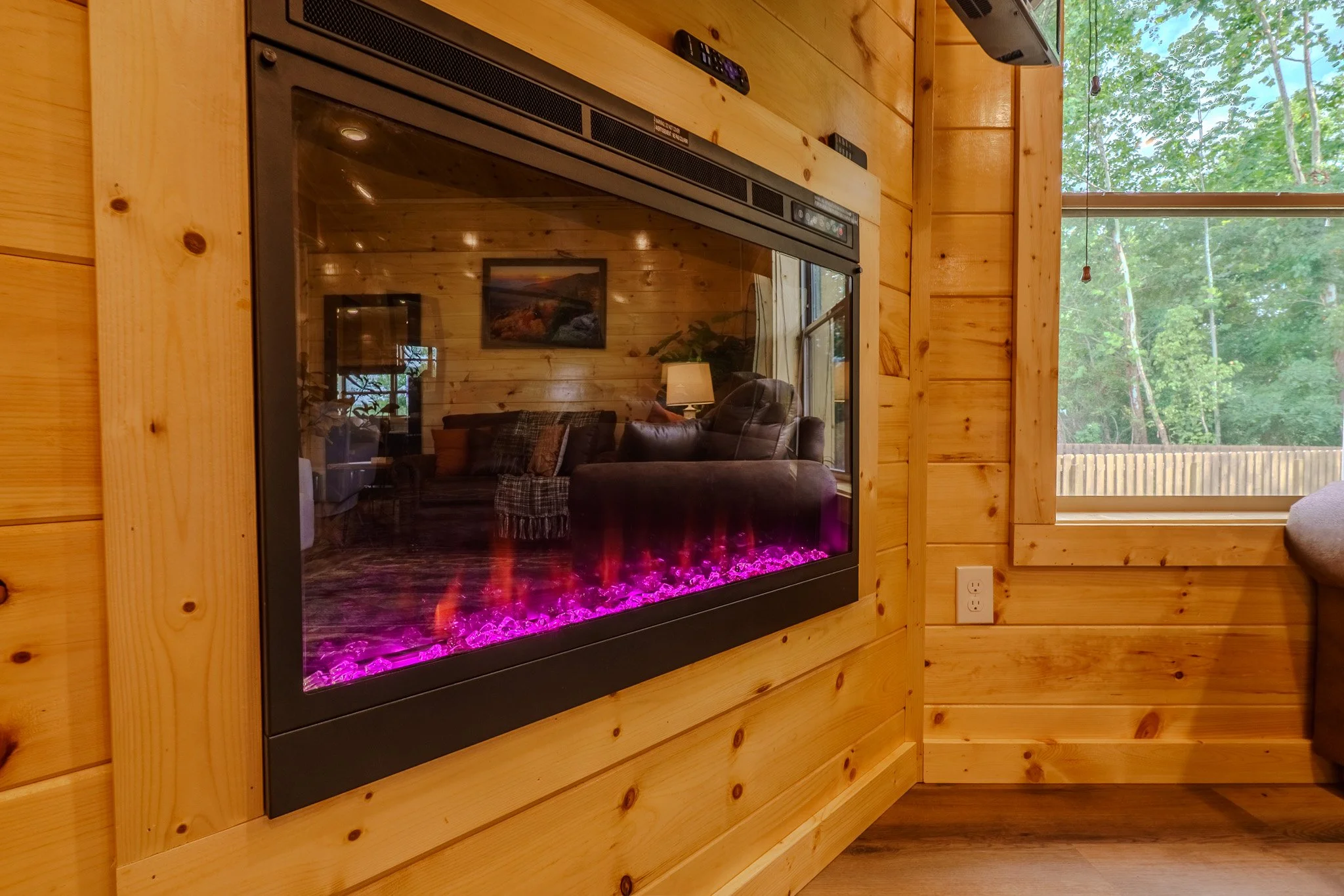 Living room with a built-in electric fireplace with purple flames, wooden walls, and a window showing green trees outside.