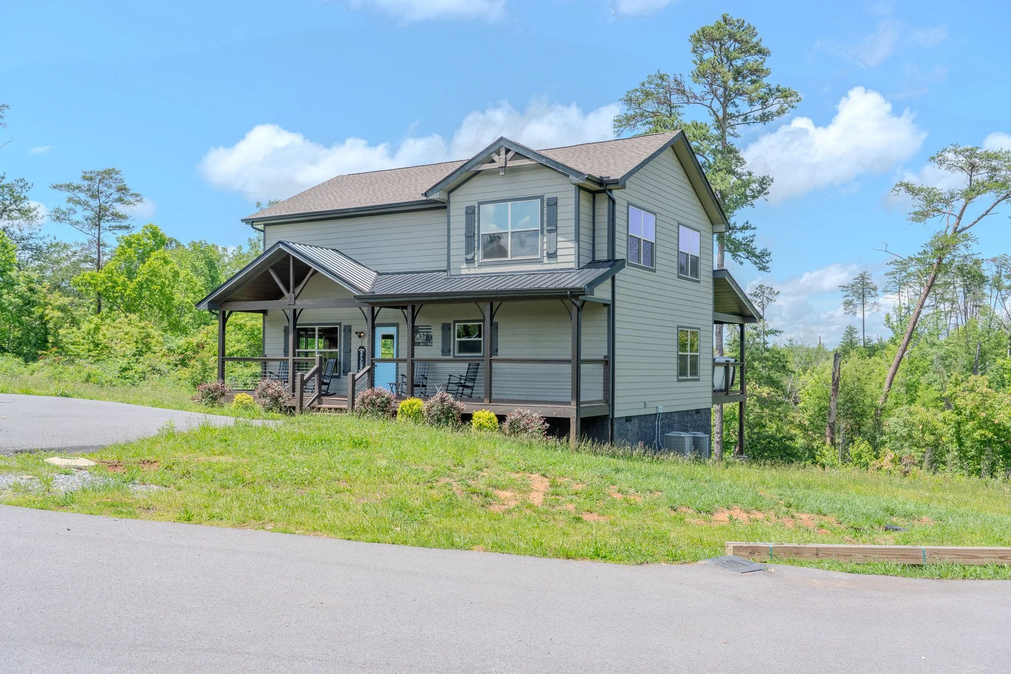 A two-story house with light gray siding and a dark gray roof, featuring a front porch with wooden railings and rocking chairs, is situated on a grassy slope with green trees and a blue sky in the background.