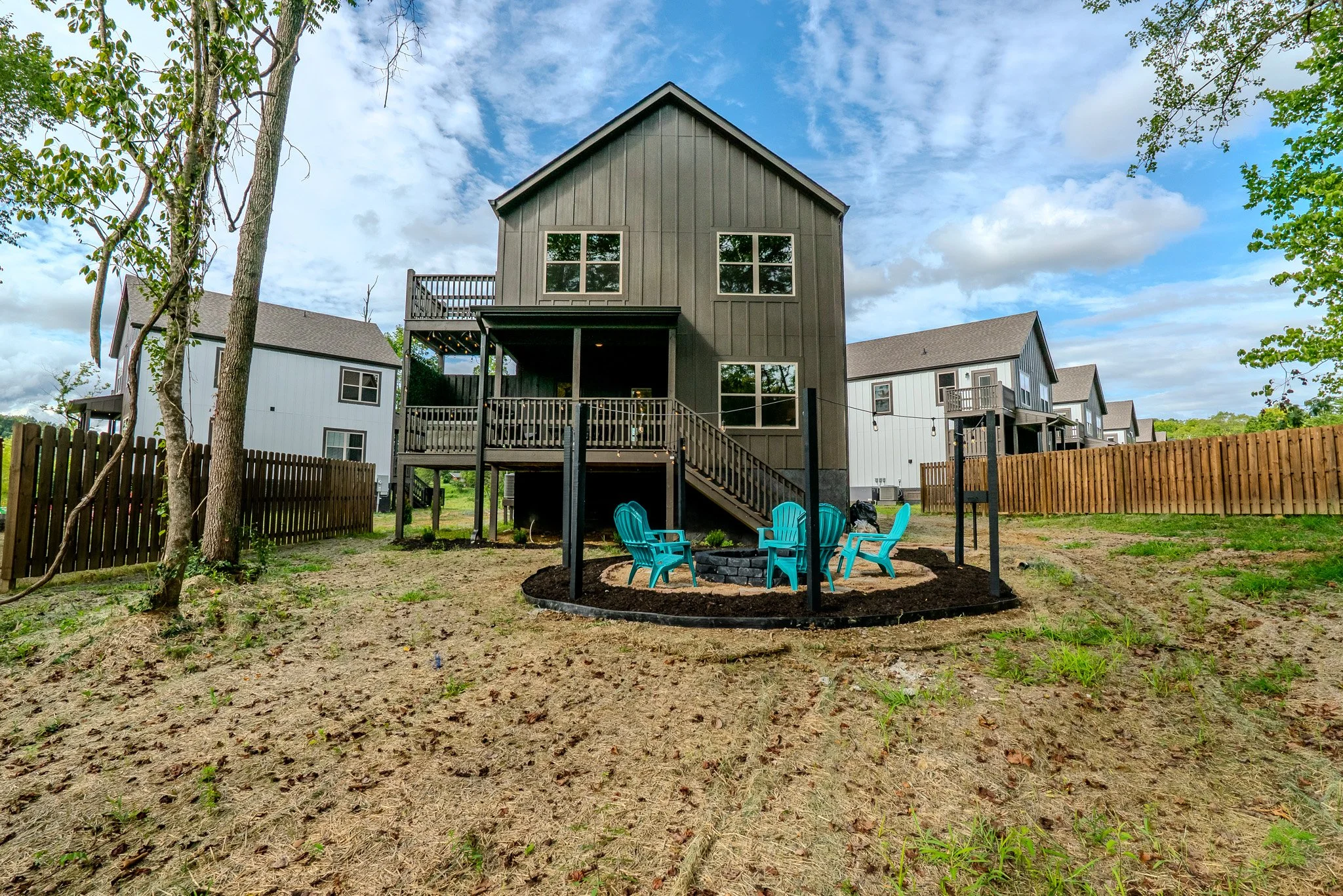 Backyard with a fire pit area surrounded by four blue Adirondack chairs and a black border, elevated wooden deck attached to a gray house, neighboring white houses, fenced yard, partly cloudy sky.