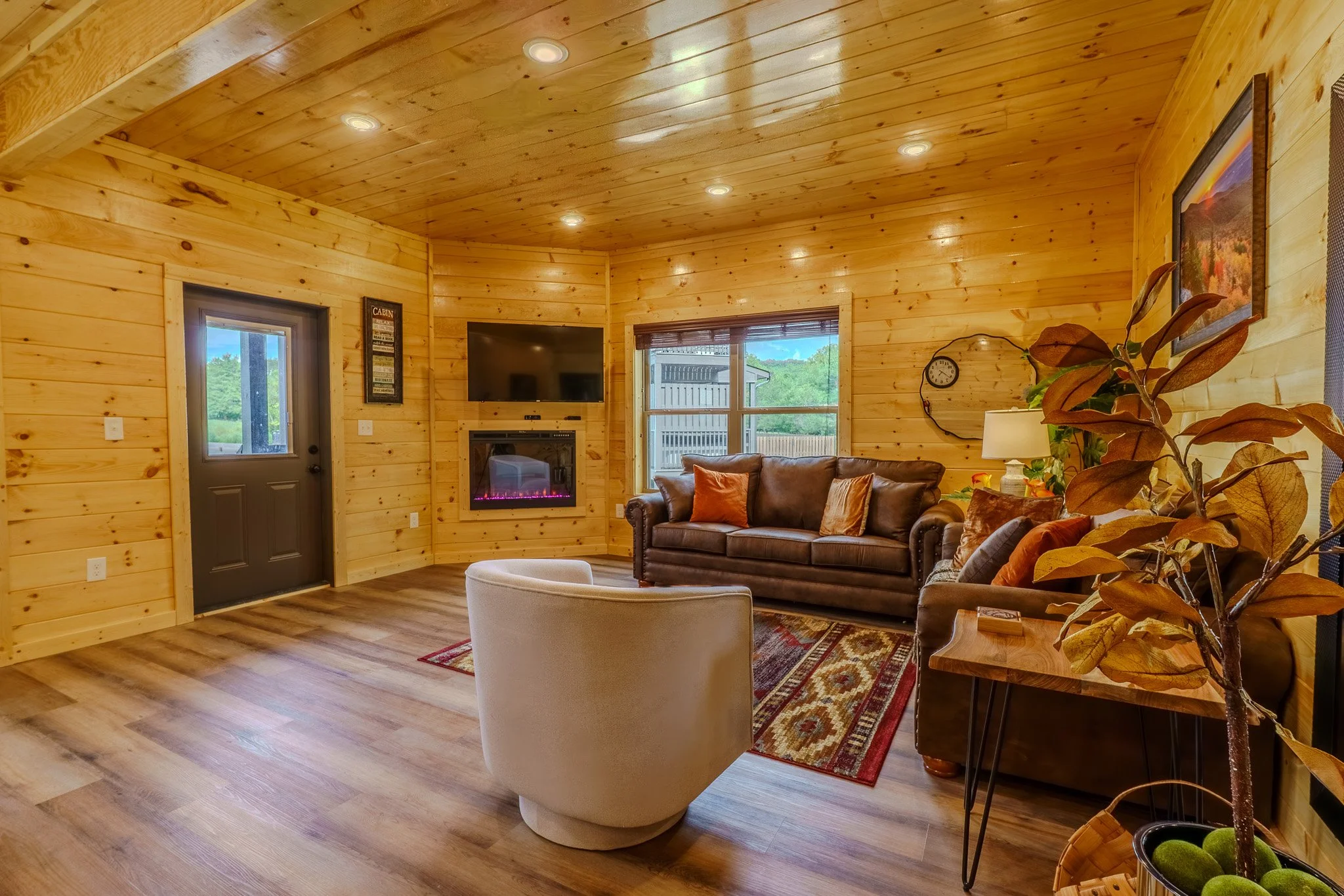 Cozy living room with wooden walls and ceiling, featuring a brown leather sofa with cushions, a white armchair, a fireplace with a TV above, a window showing outdoor greenery, a side table with a lamp, and a colorful rug.