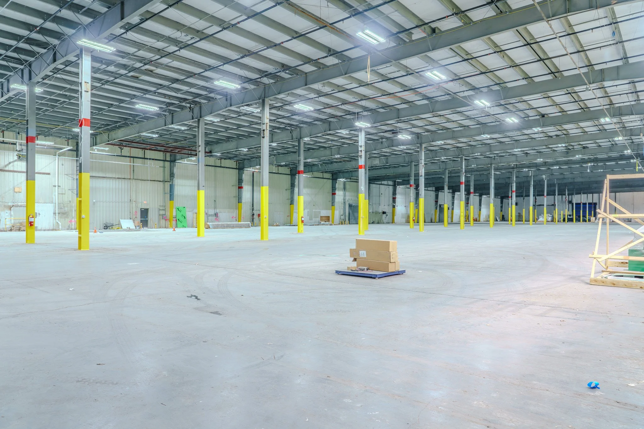 Empty warehouse with yellow safety bumpers around support columns, some cardboard boxes on a pallet, and construction materials scattered on the concrete floor.