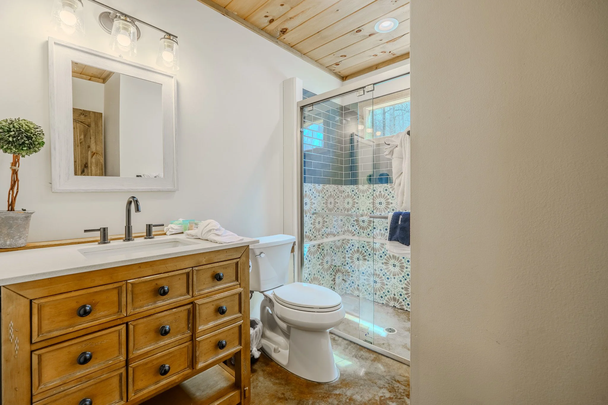Bathroom with a wooden vanity, a mirror, a white toilet, and a shower with glass door and patterned tiles.