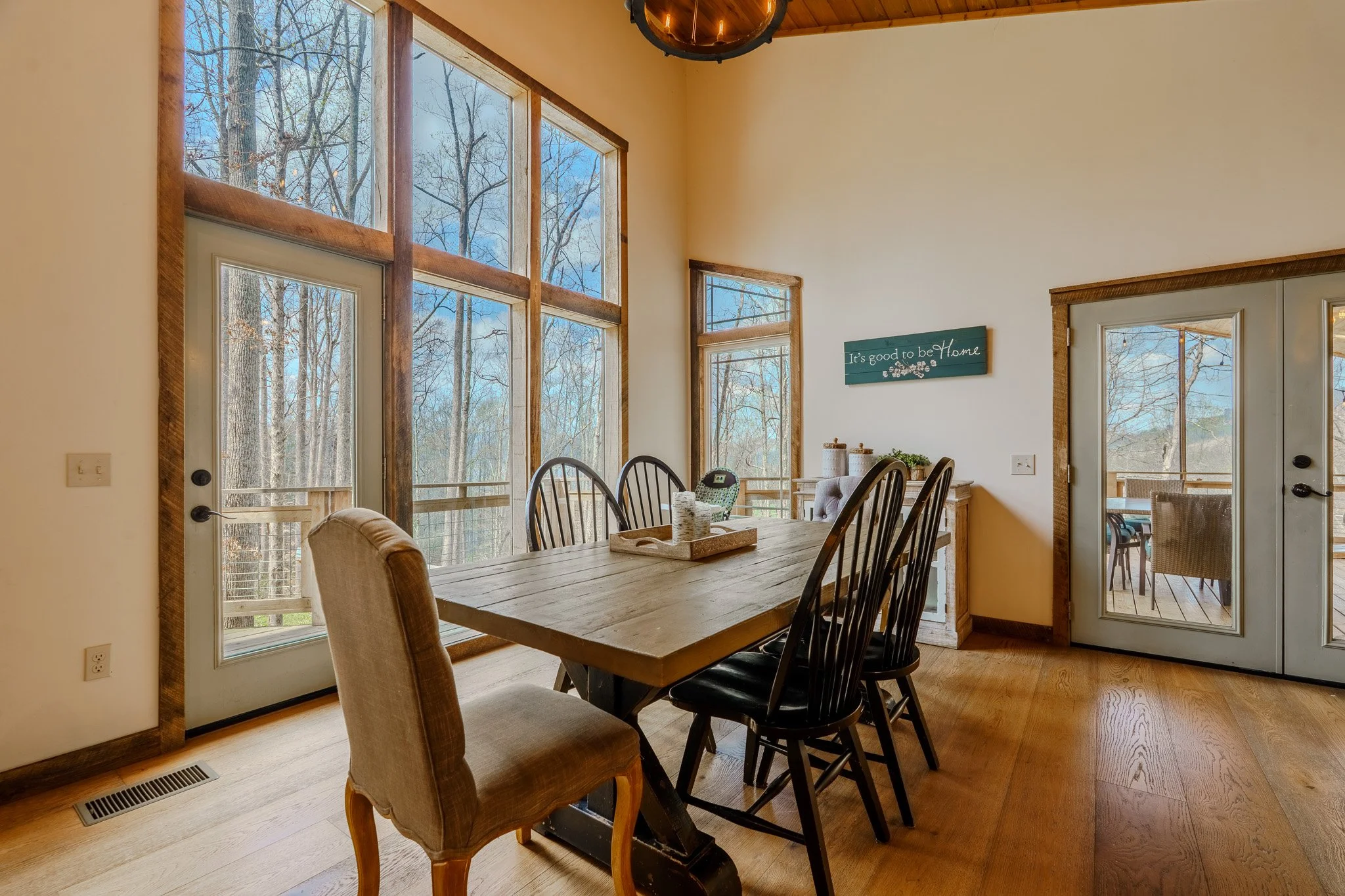 A dining room with large windows showing a wooded outdoor view, a wooden dining table with six chairs, and a sliding glass door leading outside. There is a wall sign that says 'It's good to be Home' and a small sideboard near the wall.