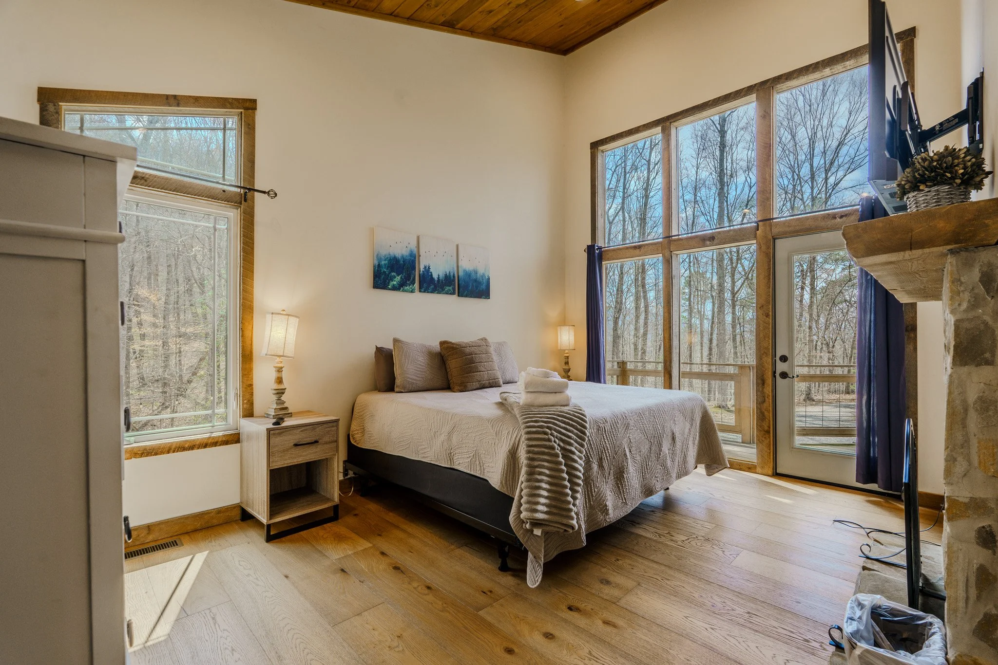 Cozy bedroom with large windows showing trees outside, a bed with folded towels, nightstands with lamps, and a mounted TV next to a stone fireplace.