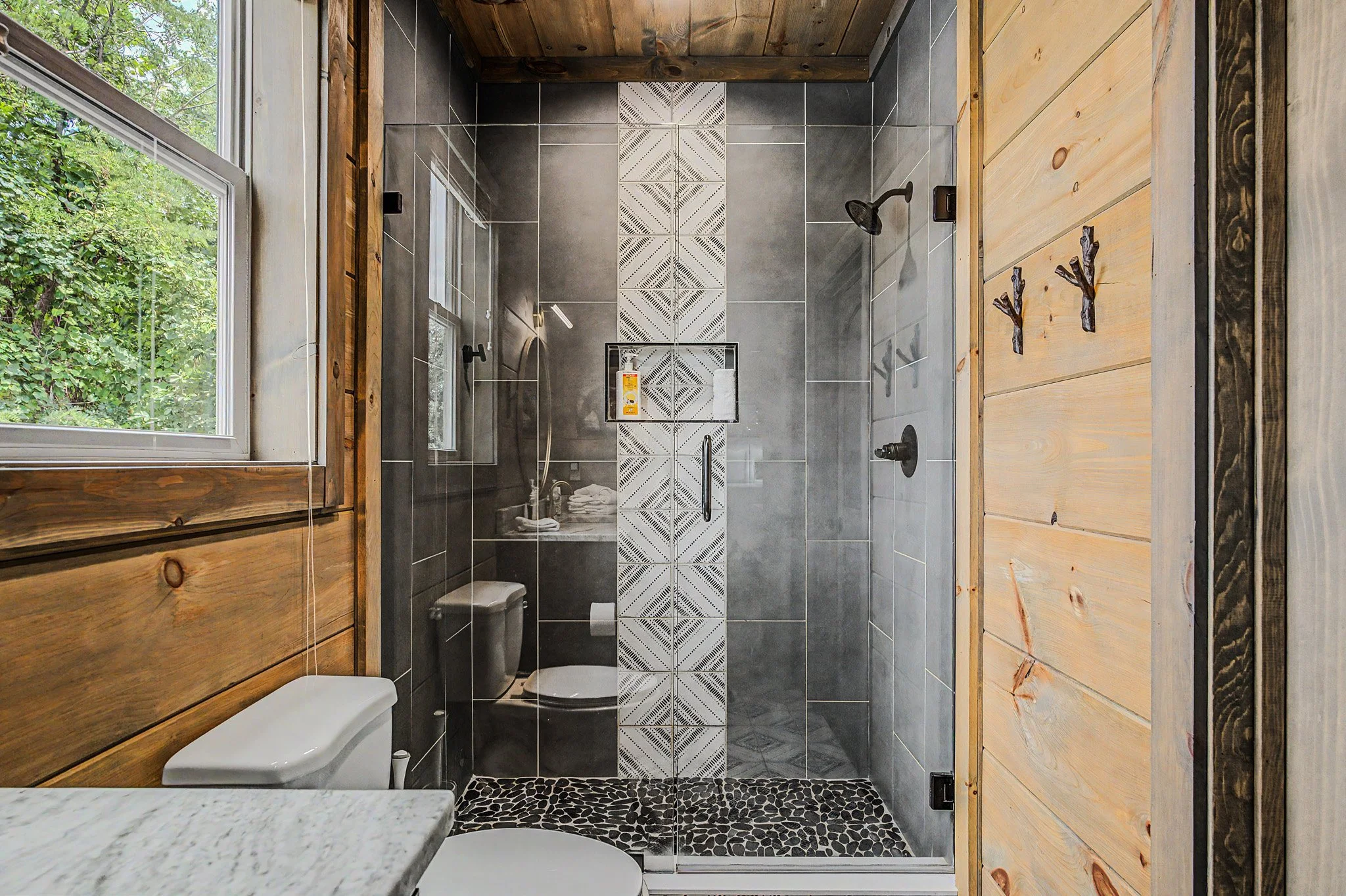 Bathroom shower with dark gray tiles, decorative patterned tile strip, rainfall showerhead, enclosed glass door, adjacent wooden wall, window with greenery outside.