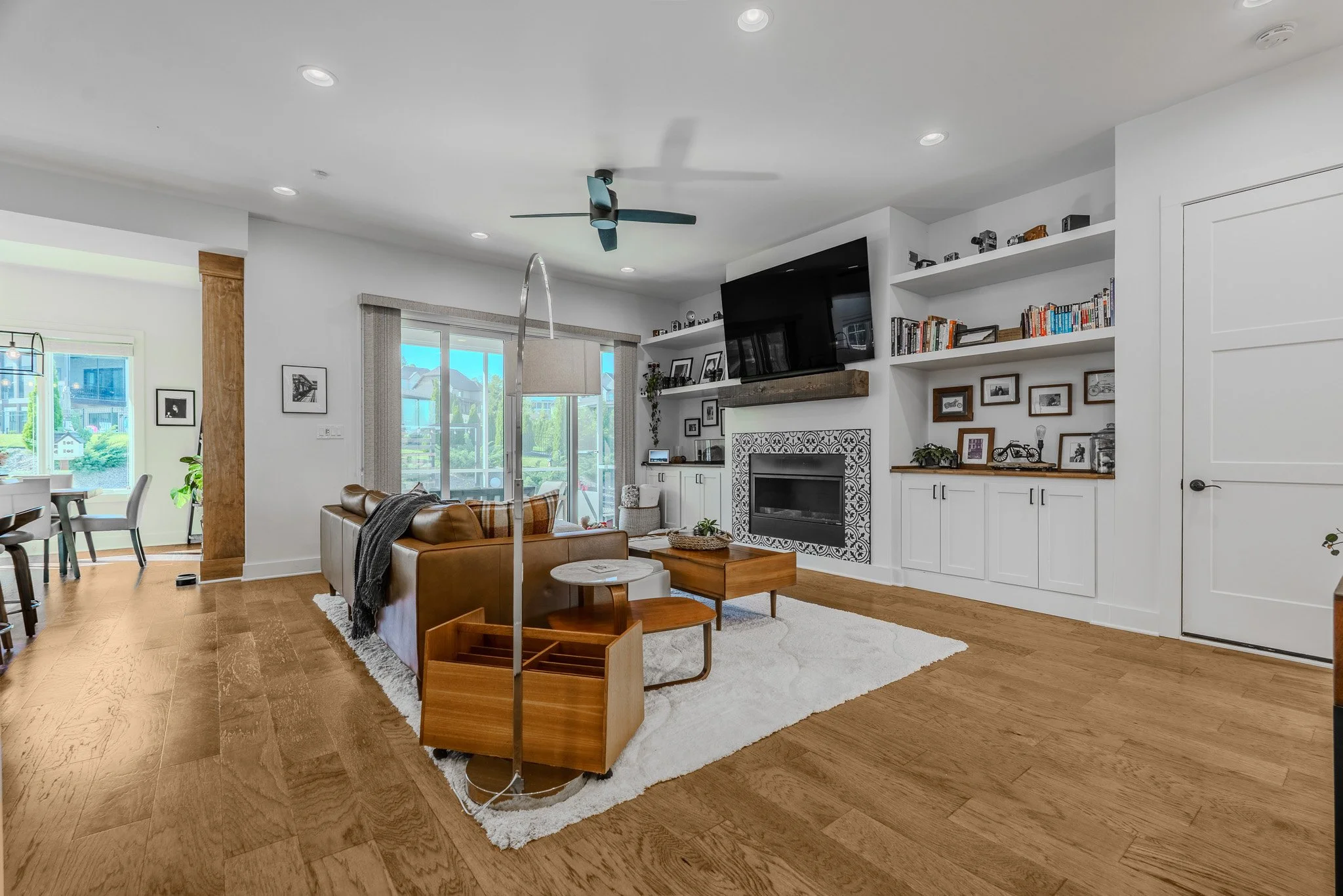 Living room with wooden floors, white walls, a white fireplace with decorative tile, a flat-screen TV above the fireplace, built-in shelves with books and photos, a brown leather sofa, a white shag rug, a round side table, a floor lamp with a curved 