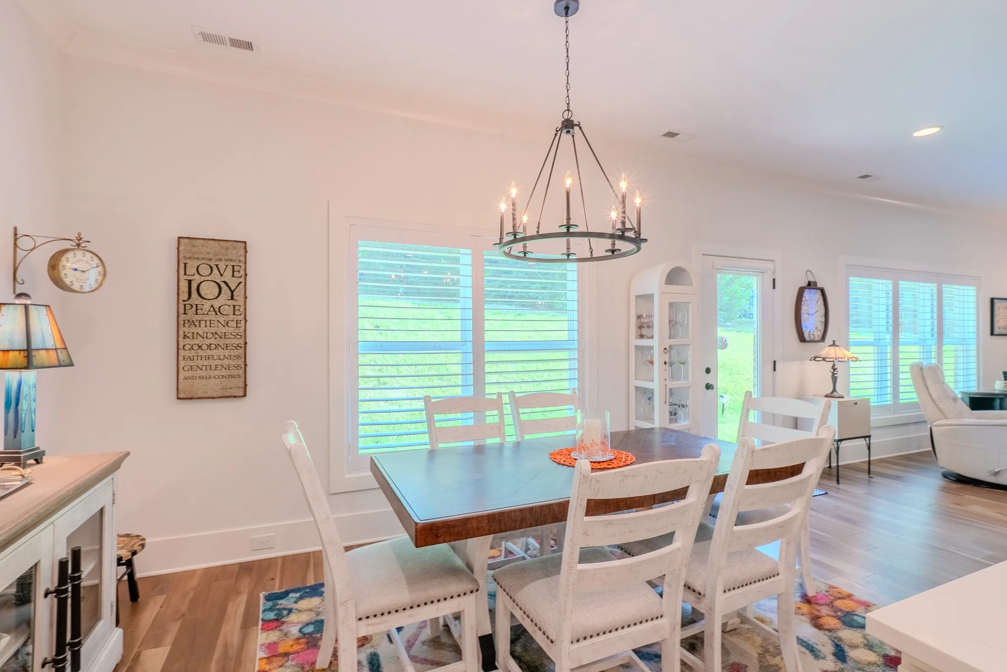 Bright dining room with wooden table and six white chairs, large windows with white blinds, chandelier, and wall decor. Part of a living area with white armchair and side table.