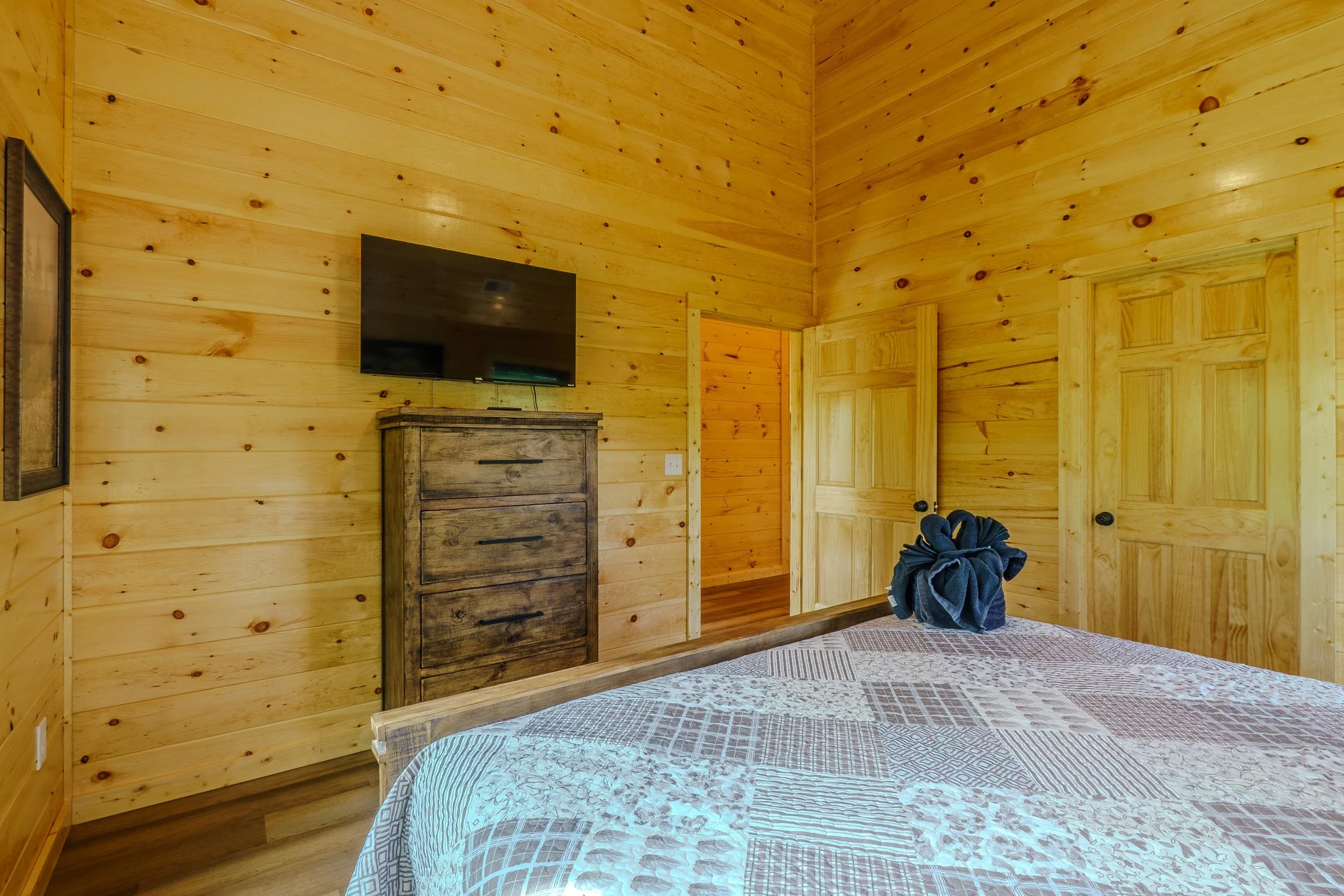 Wood-paneled bedroom with a bed, a mounted flat-screen TV, a wooden dresser, and a bundle of dark-colored clothes on the bed.