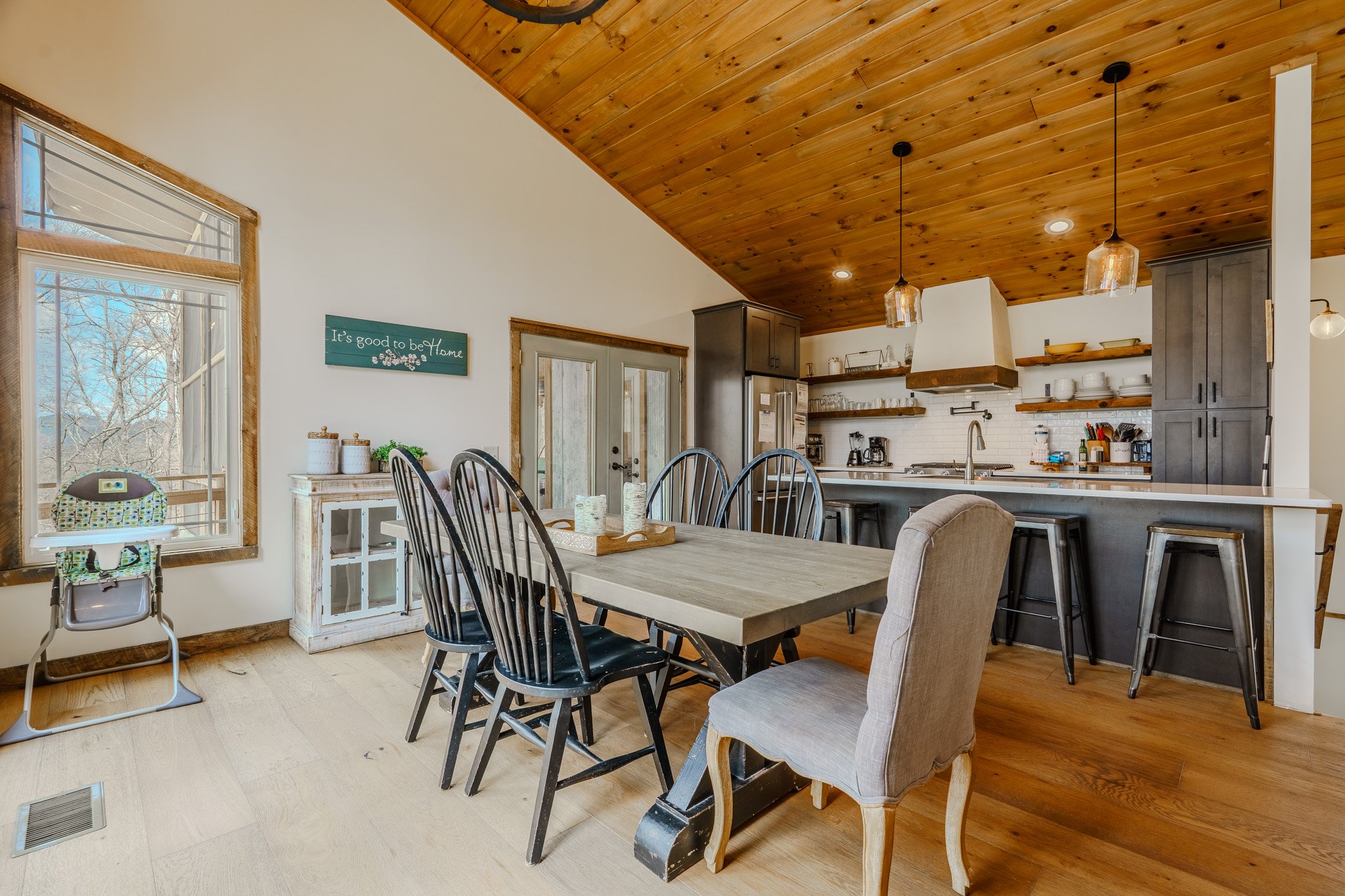 Open concept kitchen and dining area with wooden ceiling, large window, and rustic decor. Dining table with six chairs and a high chair nearby. Kitchen with black cabinets, open shelves, white backsplash, and modern appliances. Hanging pendant lights