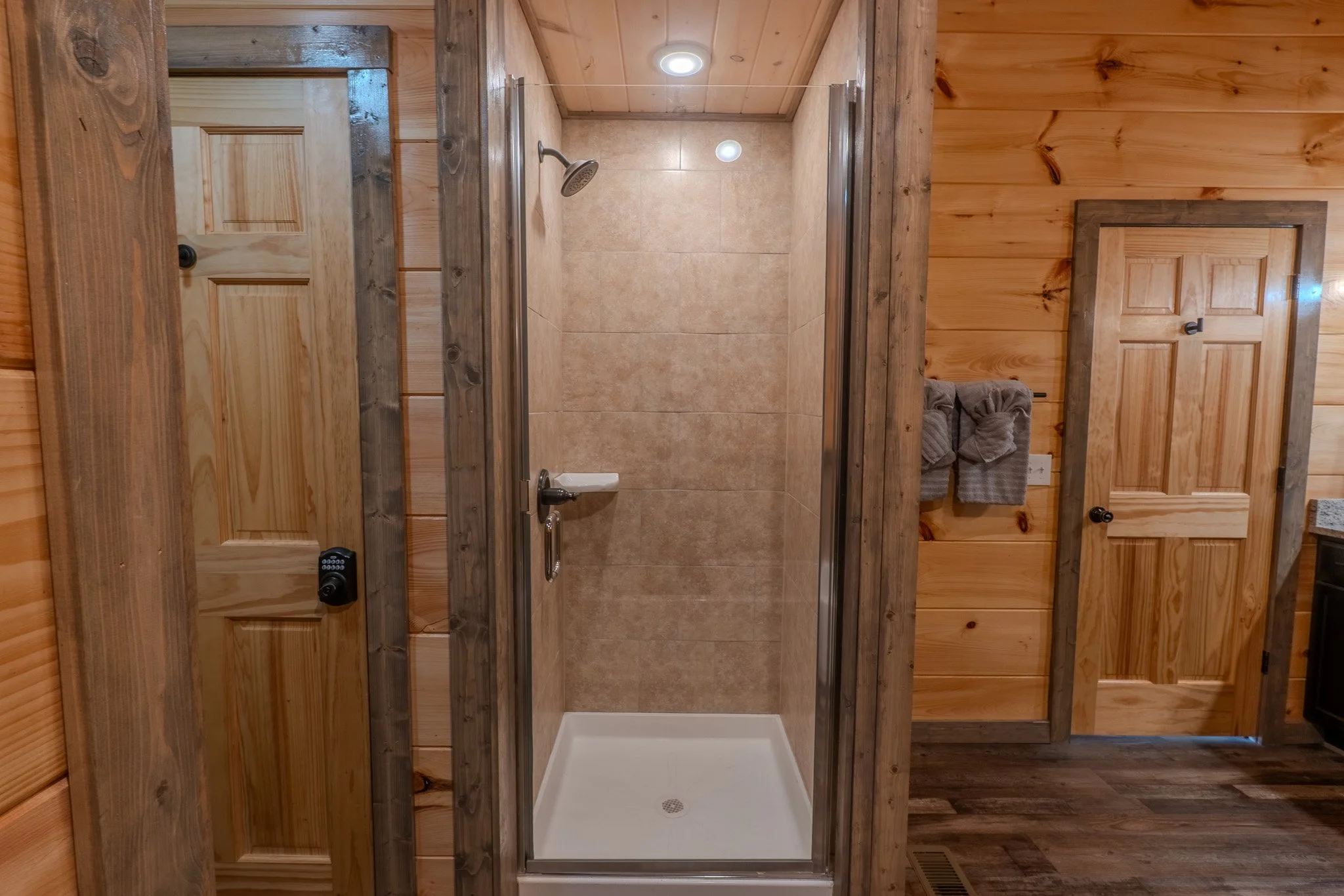 Indoor shower with wood-paneled walls, a glass door, beige tiled interior, and a small towel rack with folded towels on the right side.