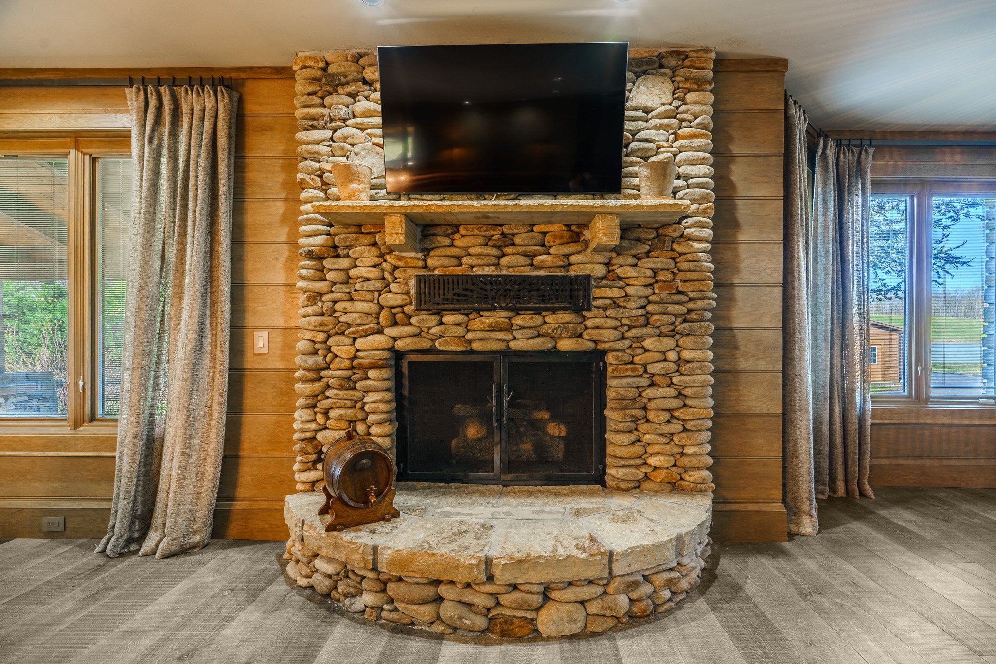 Living room with a stone fireplace, wooden walls, a flat-screen TV above the mantel, and windows with curtains on both sides.