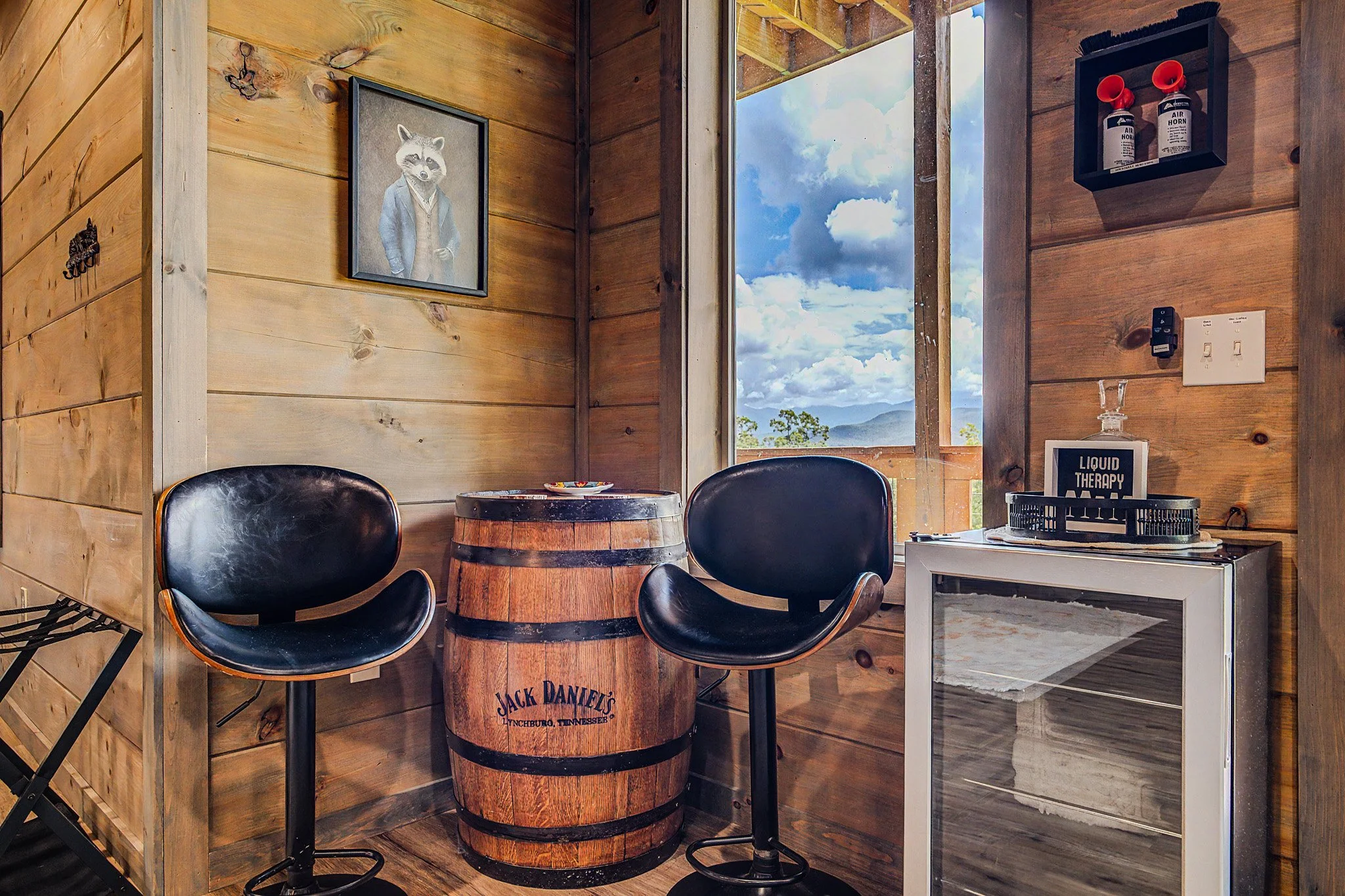 Rustic wooden interior with Jack Daniel's barrel table and black chairs, artwork of a raccoon in a jacket, window showing cloudy sky and mountains.