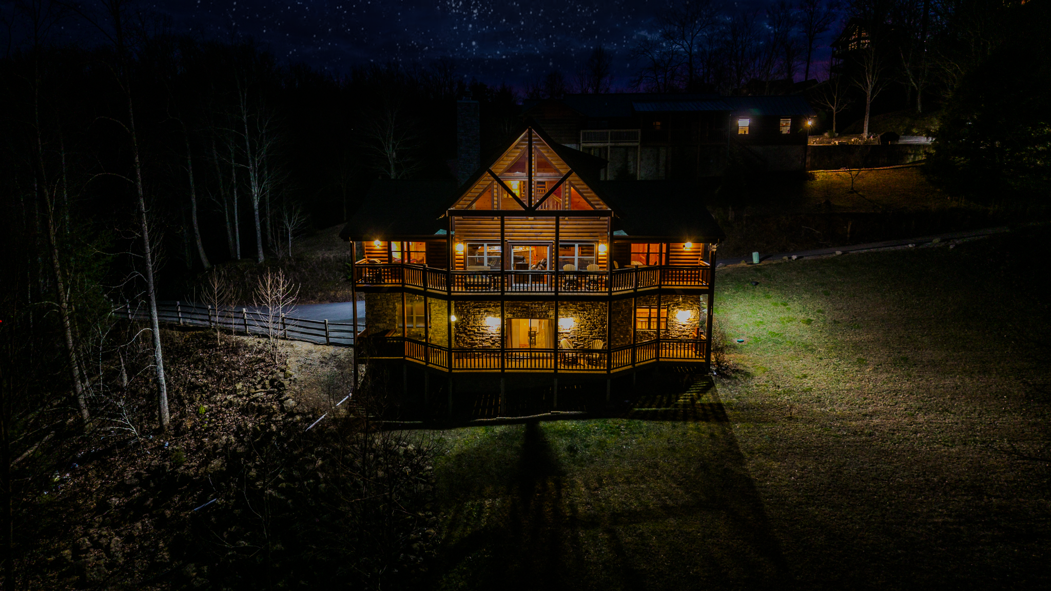 Night view of a well-lit log cabin house with large windows, surrounded by a wooded area, with a dark sky and stars above.