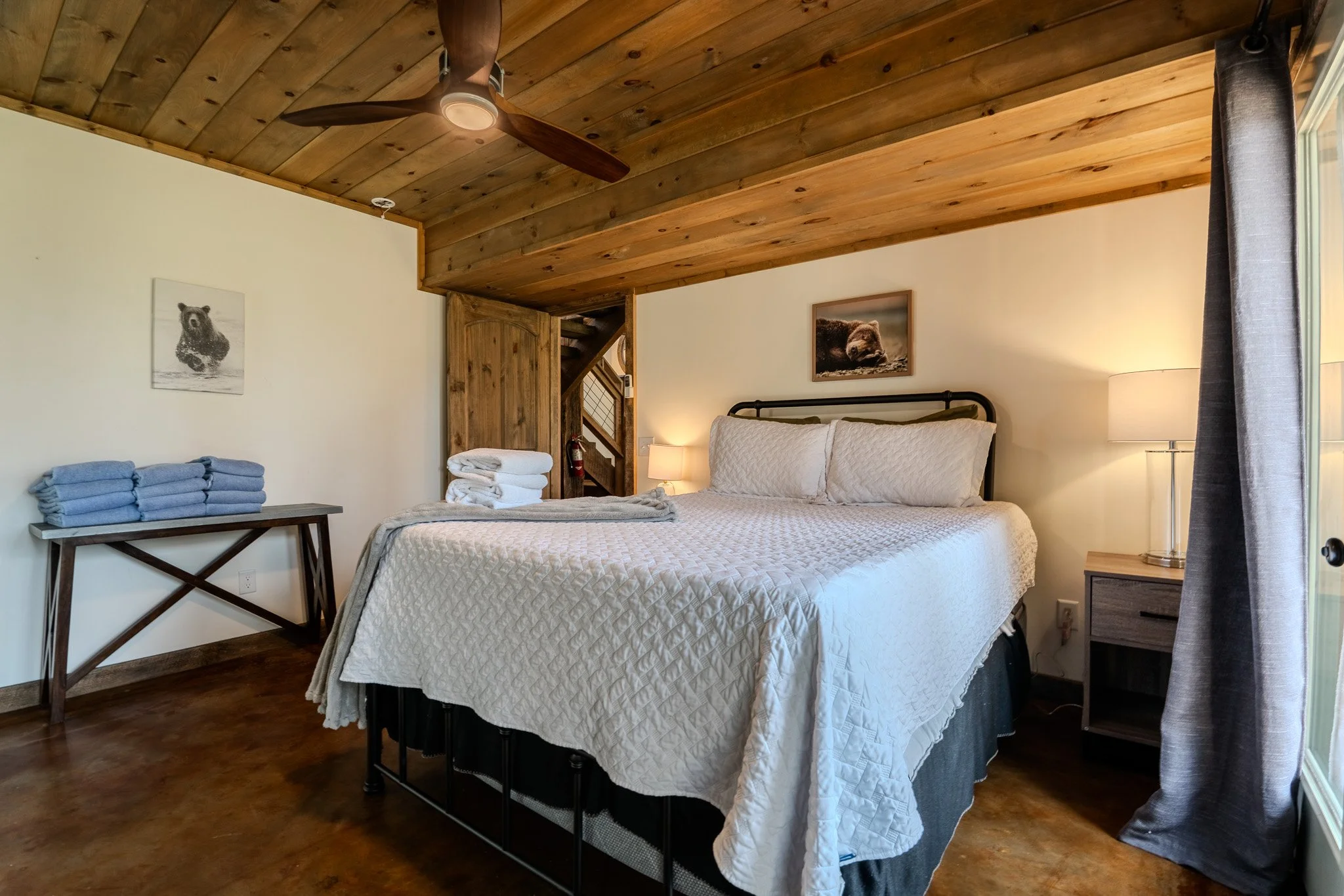 A cozy bedroom with a large bed covered in white quilted bedding, two pillows, and a picture of a bear hanging above it. There are two lamps on nightstands, and a wooden ceiling with a ceiling fan. To the left, a table with folded towels and a pictur