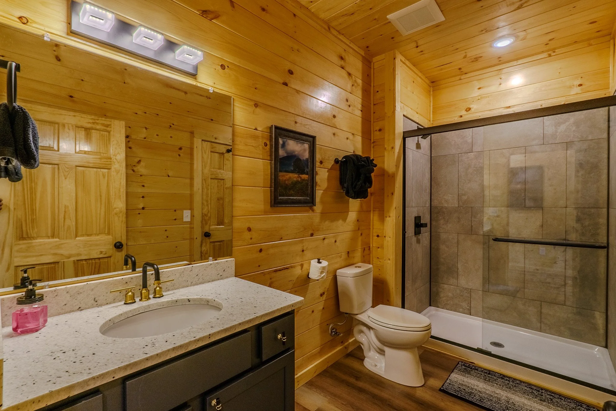 Bathroom with wood-paneled walls, a large mirror above a speckled white countertop with a sink, a black cabinet below, a toilet next to the vanity, and a glass-enclosed shower with beige tiles.