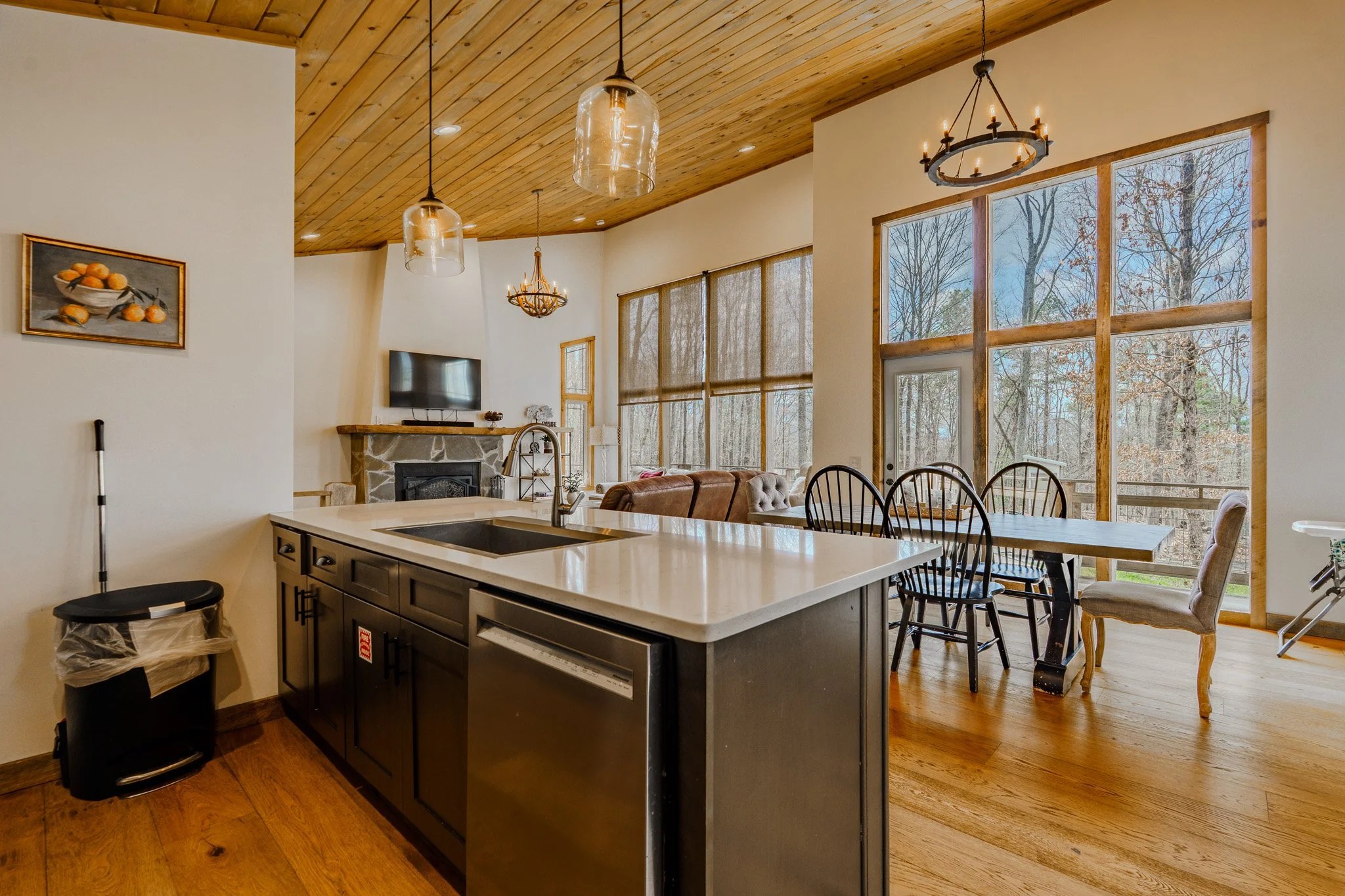 Open kitchen and dining area with wood ceiling, large windows, stone fireplace, and modern chandelier. Includes kitchen island, dining table, chairs, and a view of trees outside.