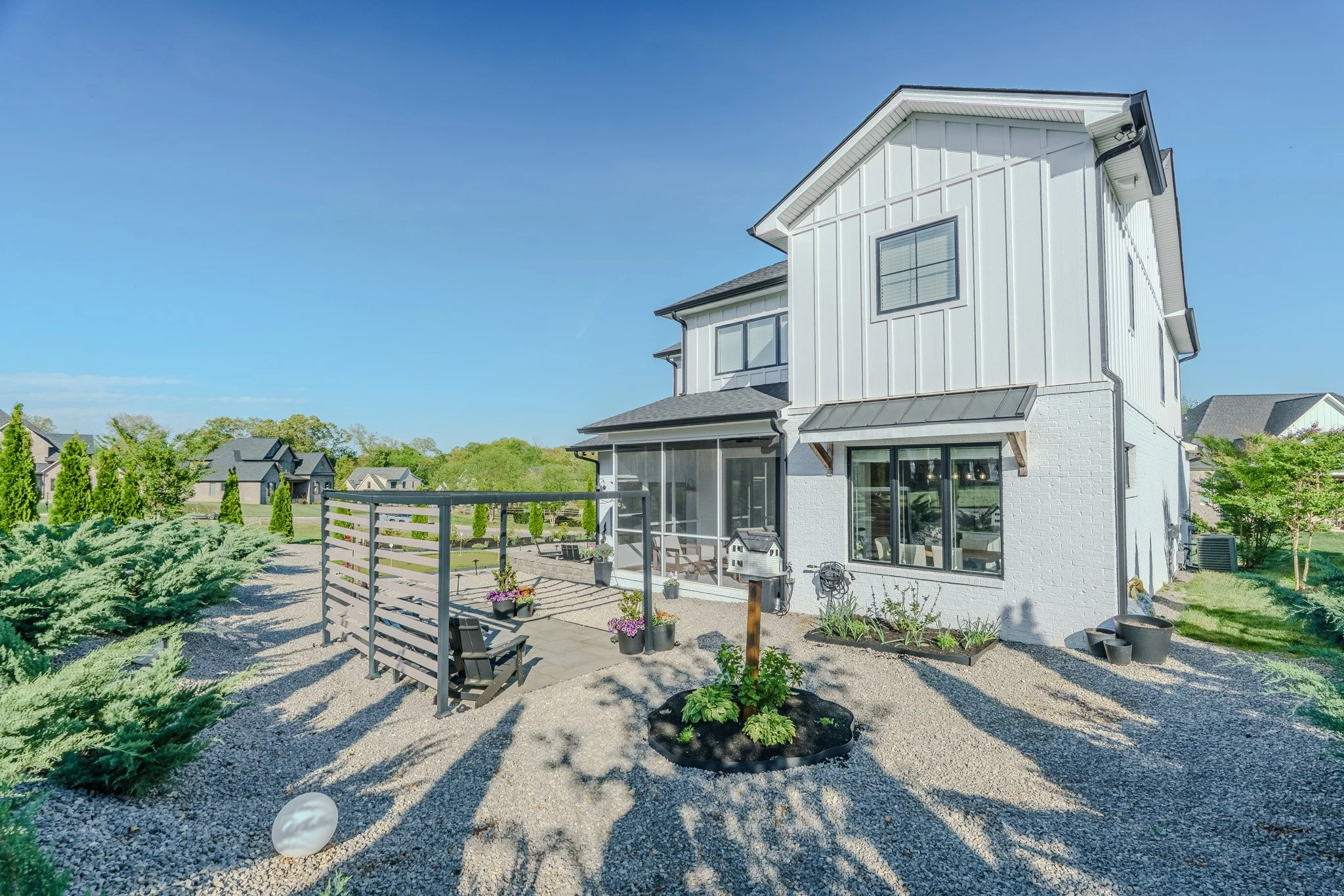 A modern white two-story house with a screened porch on a sunny day, surrounded by a landscaped gravel yard with plants, a small garden, and nearby neighboring houses.