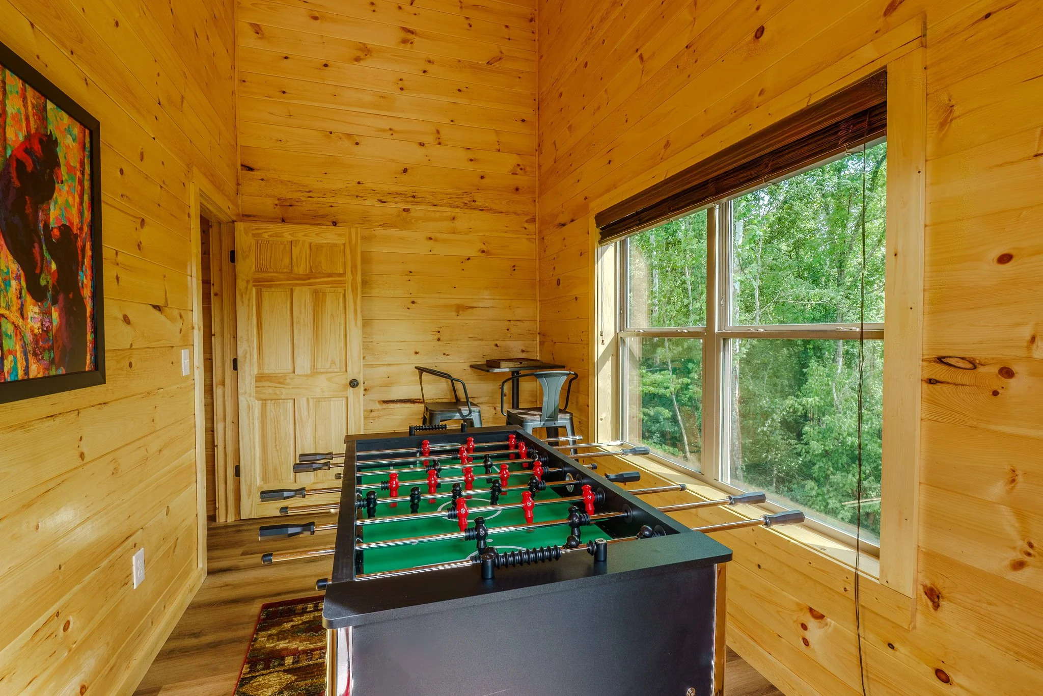 A cozy wooden game room with a foosball table near a large window showing green trees outside, and a colorful painting on the wall.