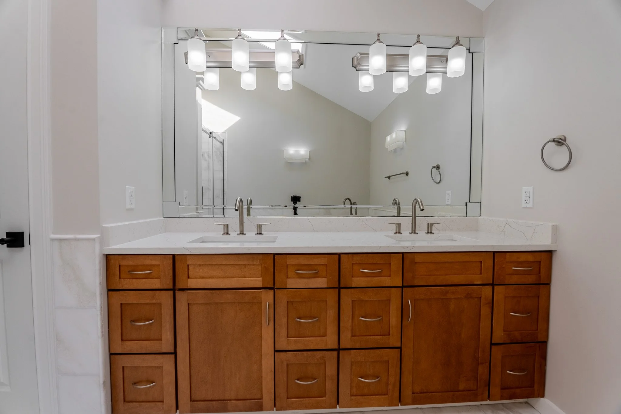 Bathroom vanity with a large mirror, wooden cabinets, marble countertop, and dual sinks with modern faucets, illuminated by multiple light fixtures.