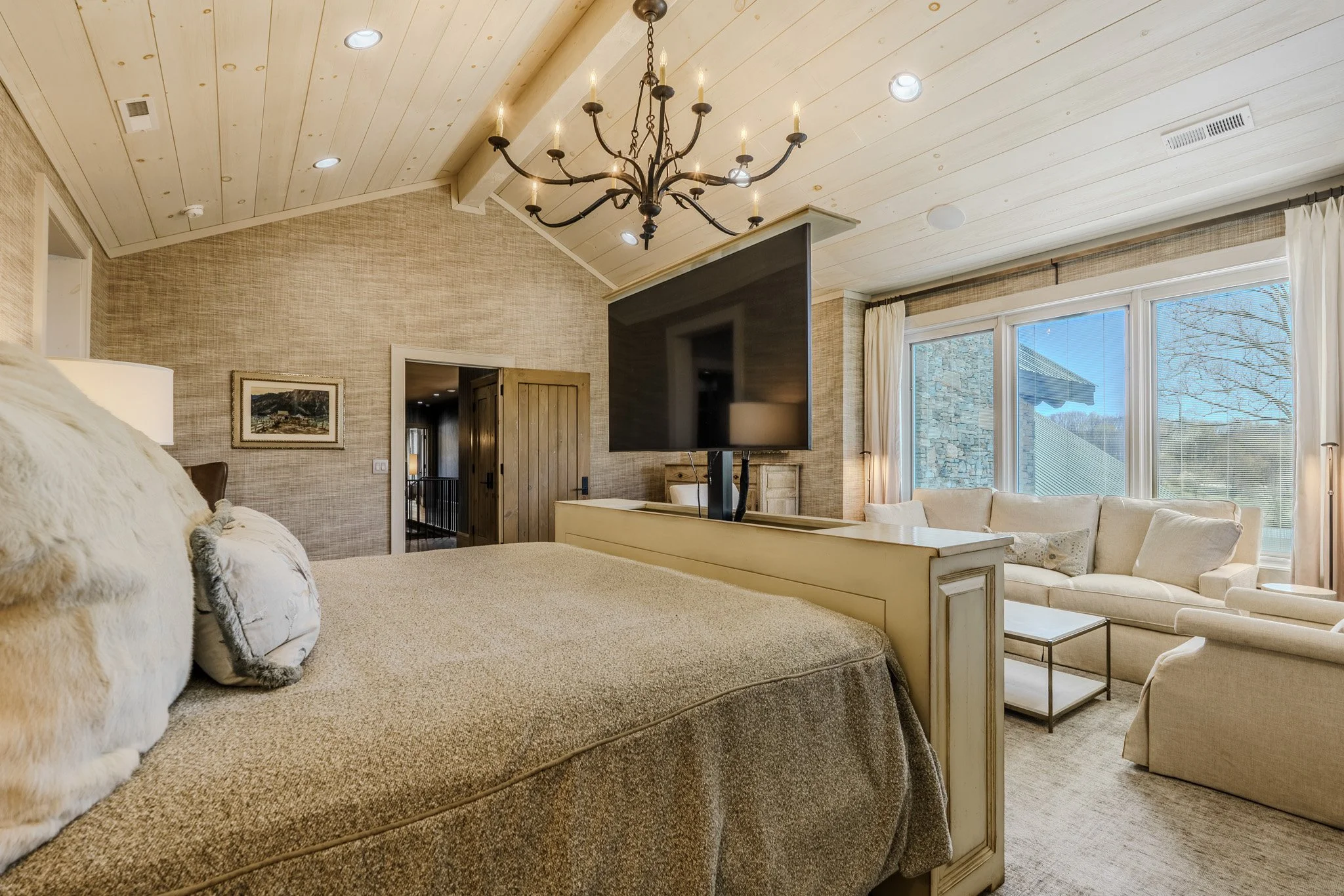 A cozy bedroom with a high, vaulted ceiling, a large bed with pillows, a chandelier hanging from the ceiling, a television mounted on a partition, a window with a view of a stone building and trees, and a sofa and coffee table near the window.