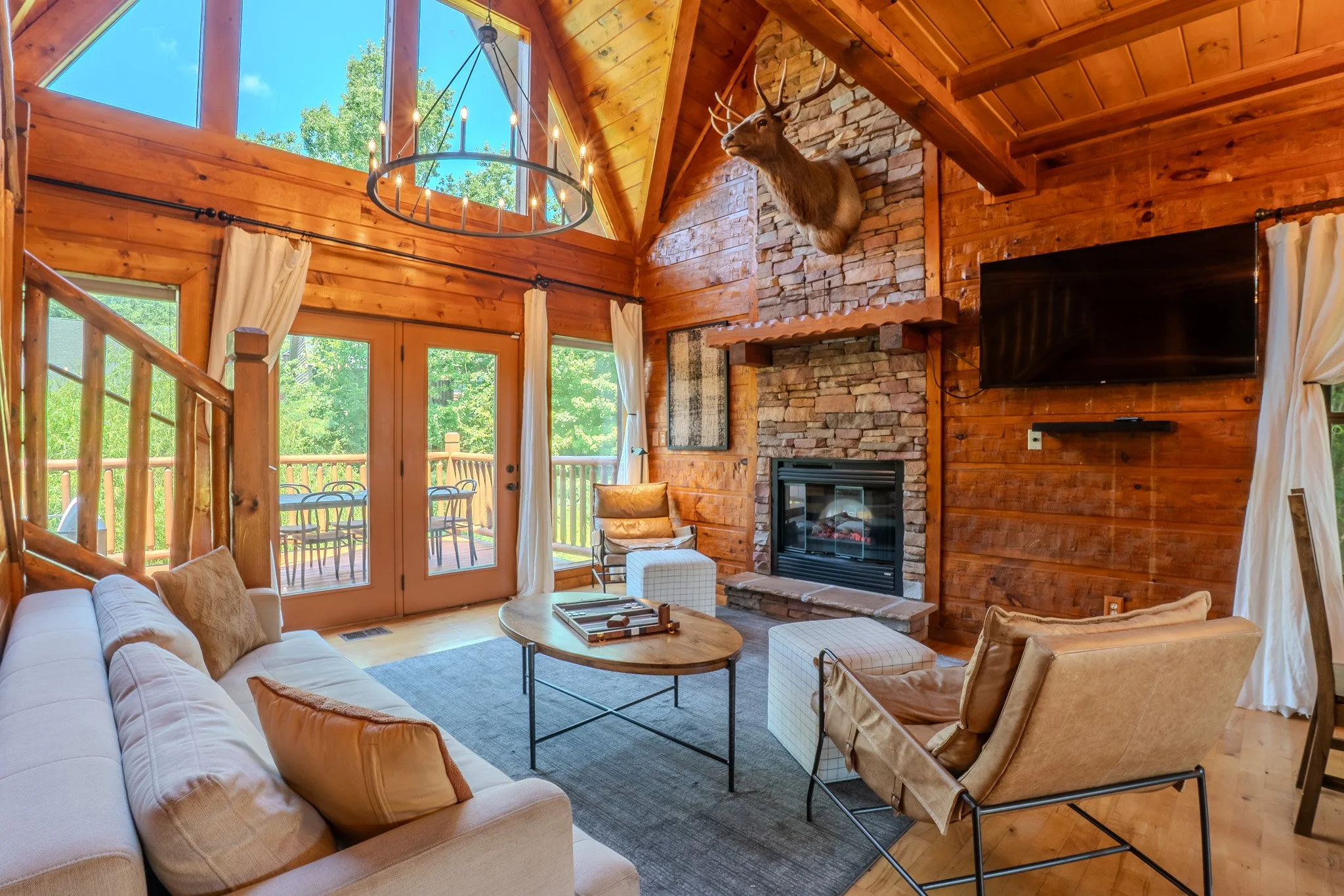 Living room with wooden walls and ceiling, large windows, and a glass door leading to a deck. A mounted deer head is above a stone fireplace. There is a flat-screen TV on the wall, and furniture includes a white sofa, an armchair, a round coffee tabl