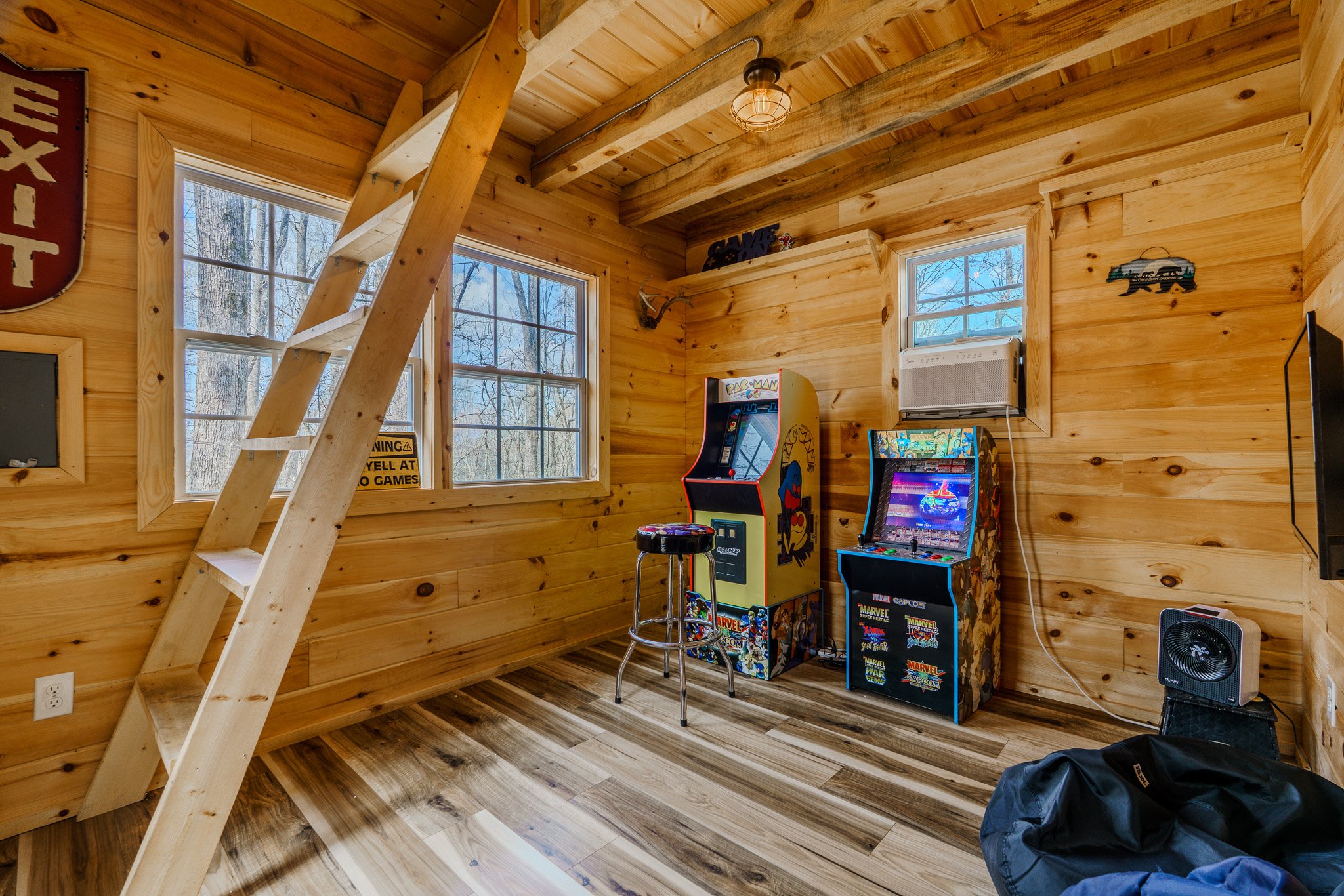 Wood-paneled room with two arcade game machines, a stool, a window air conditioning unit, a small speaker, and a ladder leading to a loft.