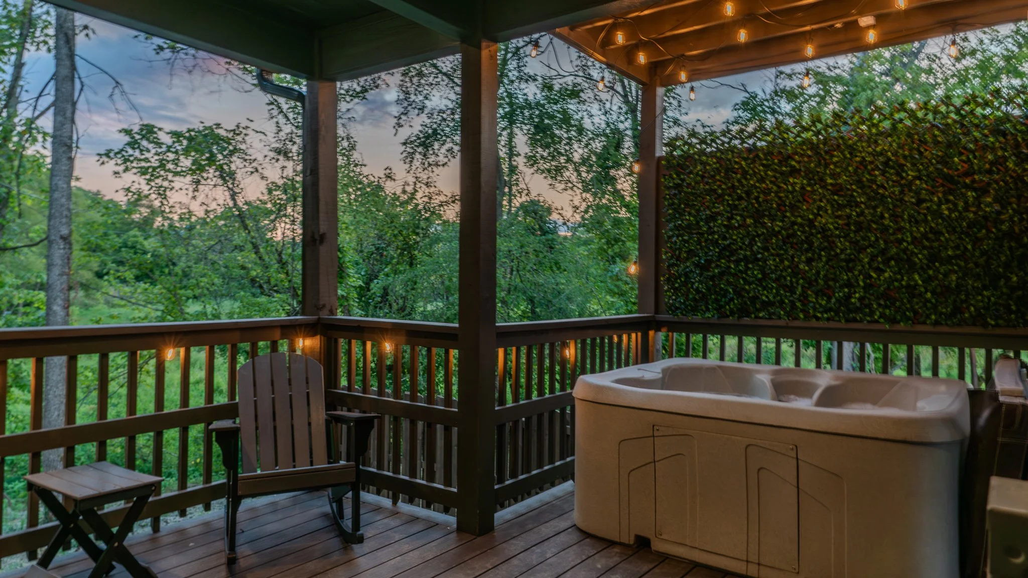 Cozy outdoor deck with hot tub, Adirondack chair, small table, string lights, surrounded by green trees and foliage, during dusk.