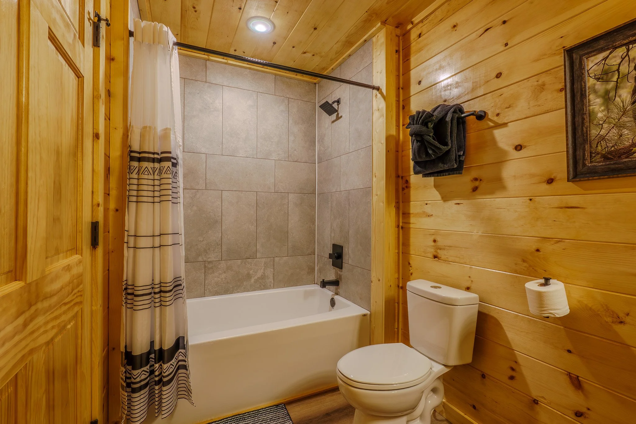Bathroom with wood-paneled walls, a bathtub with a shower curtain, a toilet, a towel rack with dark towels, a framed picture, and a toilet paper holder.