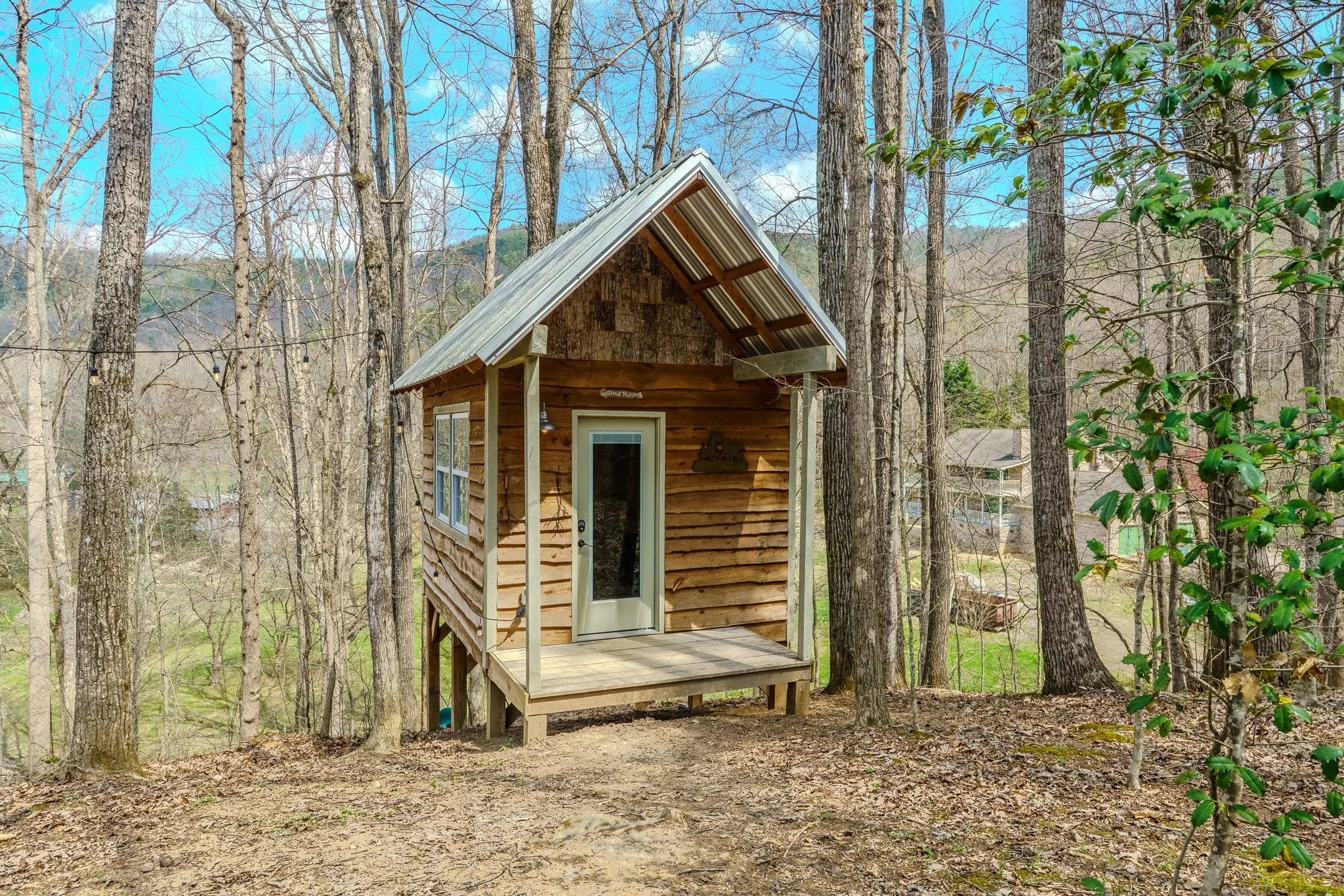 Small wooden cabin with a porch, situated in a wooded area with tall trees and a clear blue sky in the background.