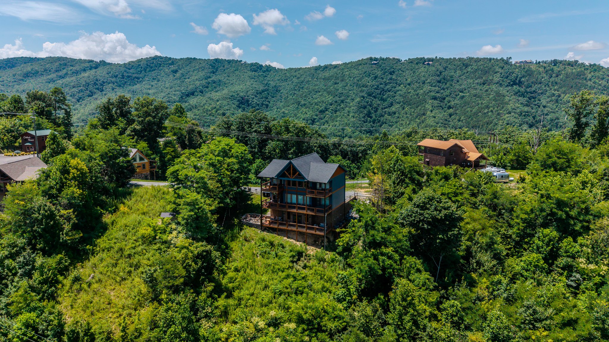 A scenic view of a green, hilly landscape with trees and several houses, including a new multi-story house under construction with scaffolding and a balcony, under a partly cloudy blue sky.