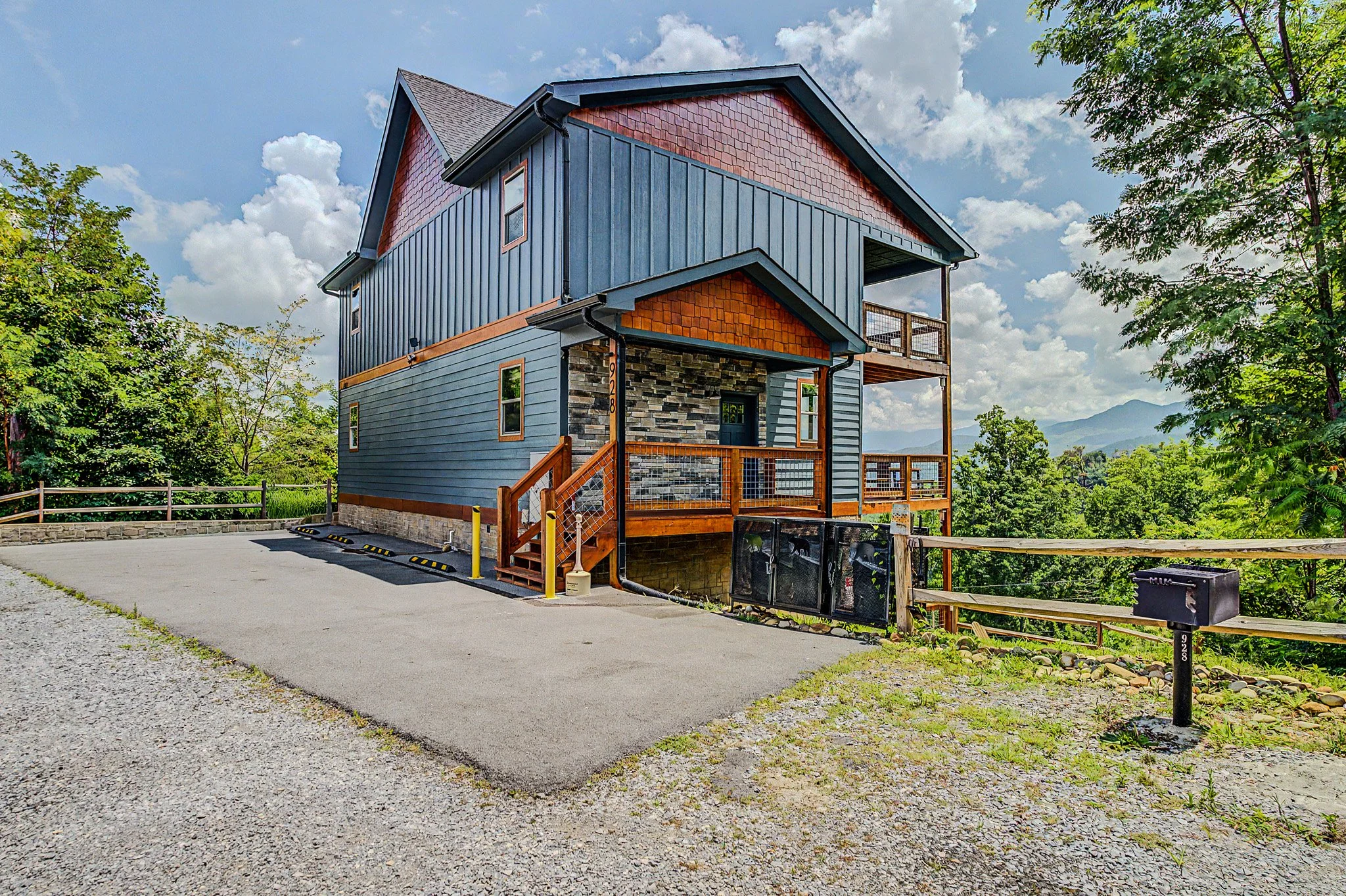 A two-story house with blue siding, stone accents, a small front porch, and a deck on the upper level, surrounded by trees and mountains in the background, under a cloudy sky.