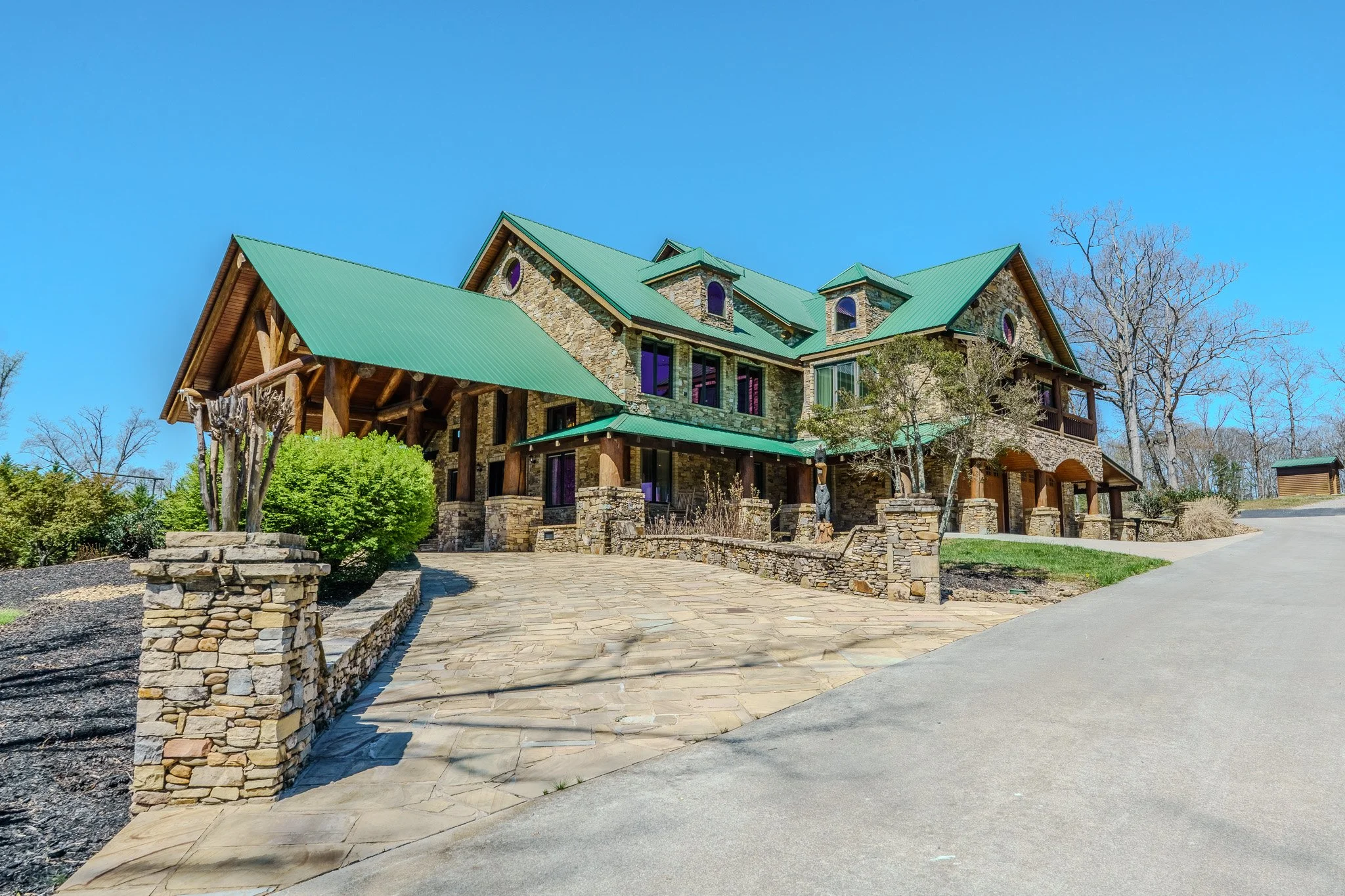 Large stone and wood house with green metal roofs and multiple levels, surrounded by trees, with a paved driveway leading up to it on a clear day.