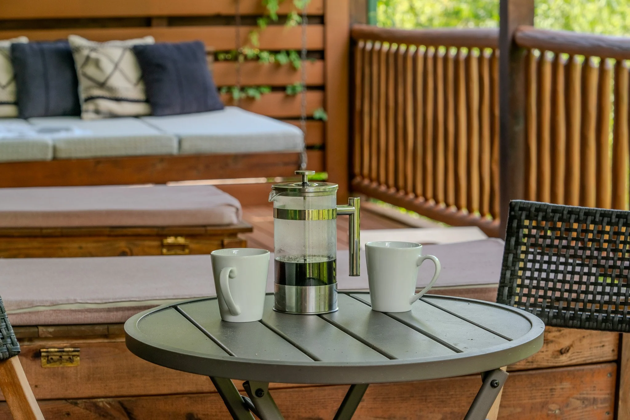 Outdoor patio table with two white coffee mugs and a French press, with a wooden bench and a cushioned lounging area in the background.