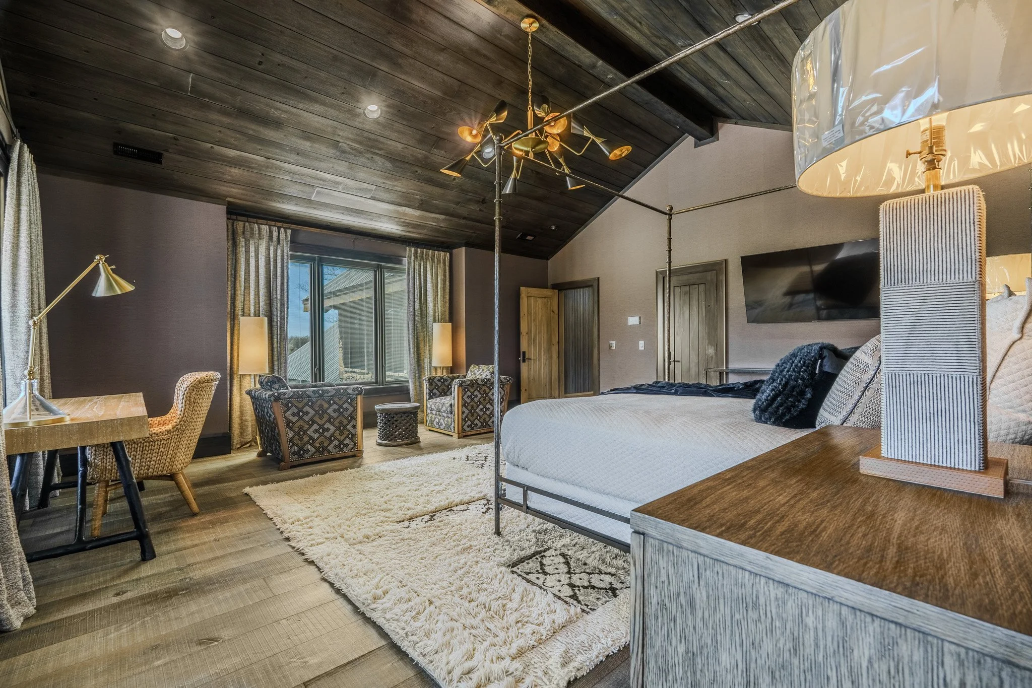 A modern bedroom with a large bed, wooden nightstand with a lamp, a flat-screen TV, a cozy sitting area with two patterned armchairs, a small round table, a window with curtains, a wooden ceiling with modern chandelier, and light-colored wooden floor