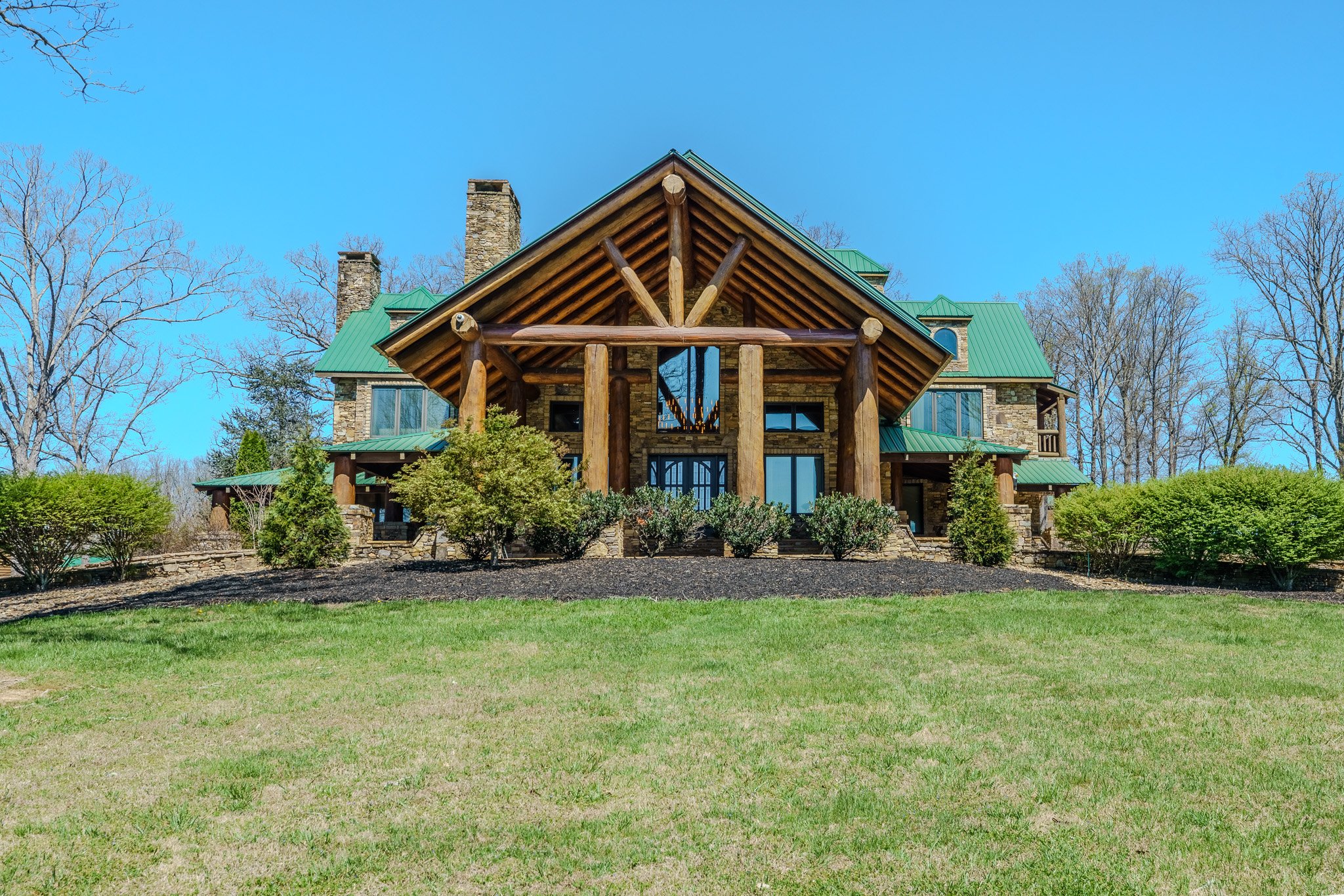 Large house with a stone exterior, green metal roof, and prominent log pillars on a lawn with trees in the background.