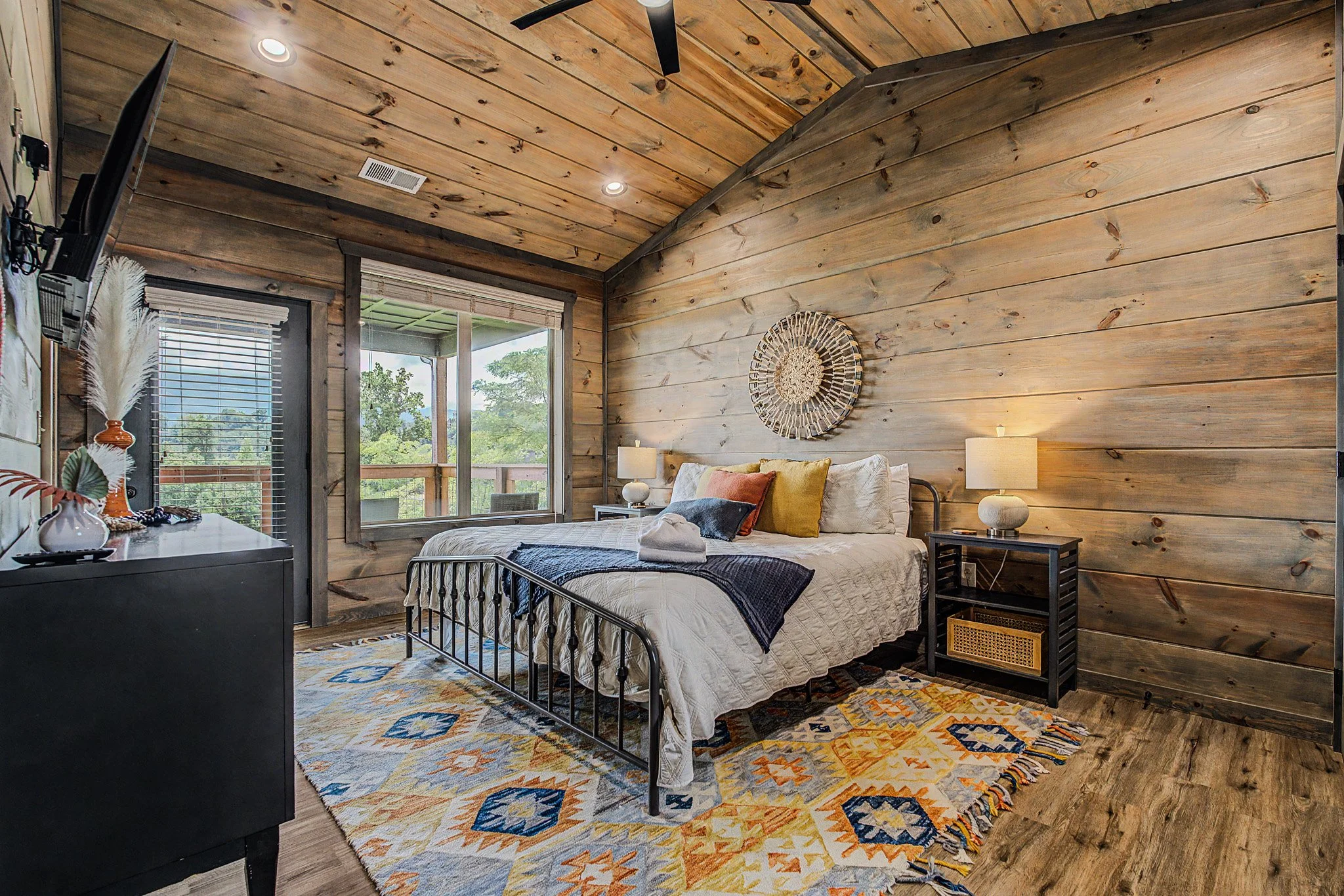 A cozy bedroom with wood-paneled walls and ceiling, a metal bed frame with white bedding and colorful pillows, a patterned area rug, a dresser with decorative objects, and a large window with a view of trees outside.