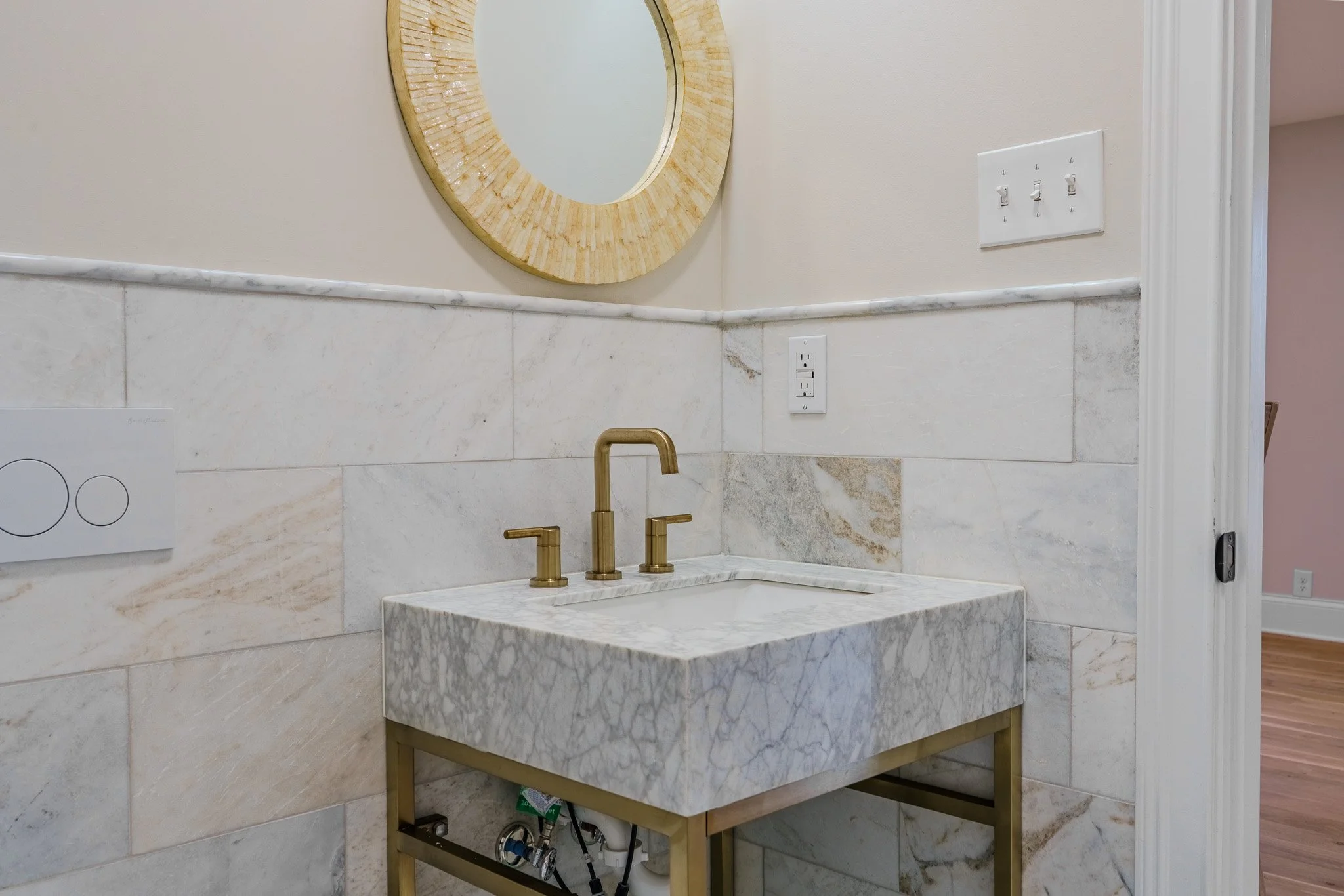 A marble bathroom sink with gold fixtures and a round mirror above it, with a beige wall and electrical outlets nearby.