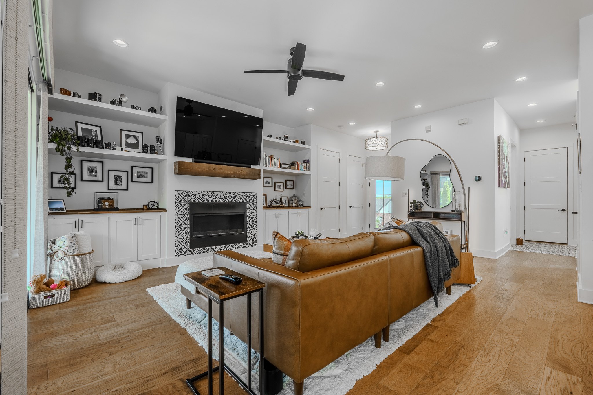 Living room with a leather sofa, wooden side table, and a white rug. Built-in shelves with books and photos, a mounted flat-screen TV, a fireplace with decorative tile, and a ceiling fan. Bright and modern decor with natural light.