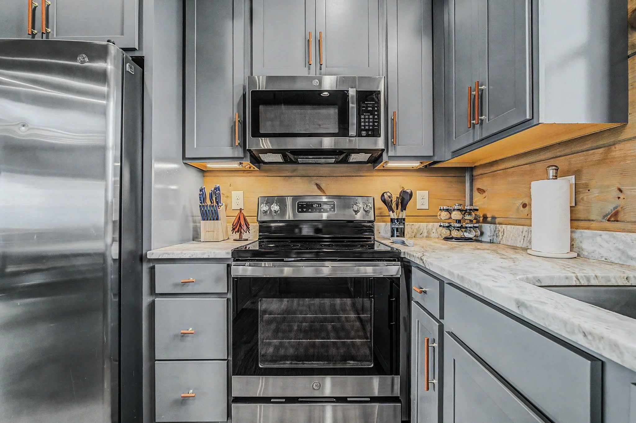 A modern kitchen with gray cabinets, a stainless steel microwave, electric stove, and a granite countertop with a paper towel roll and spice jars.