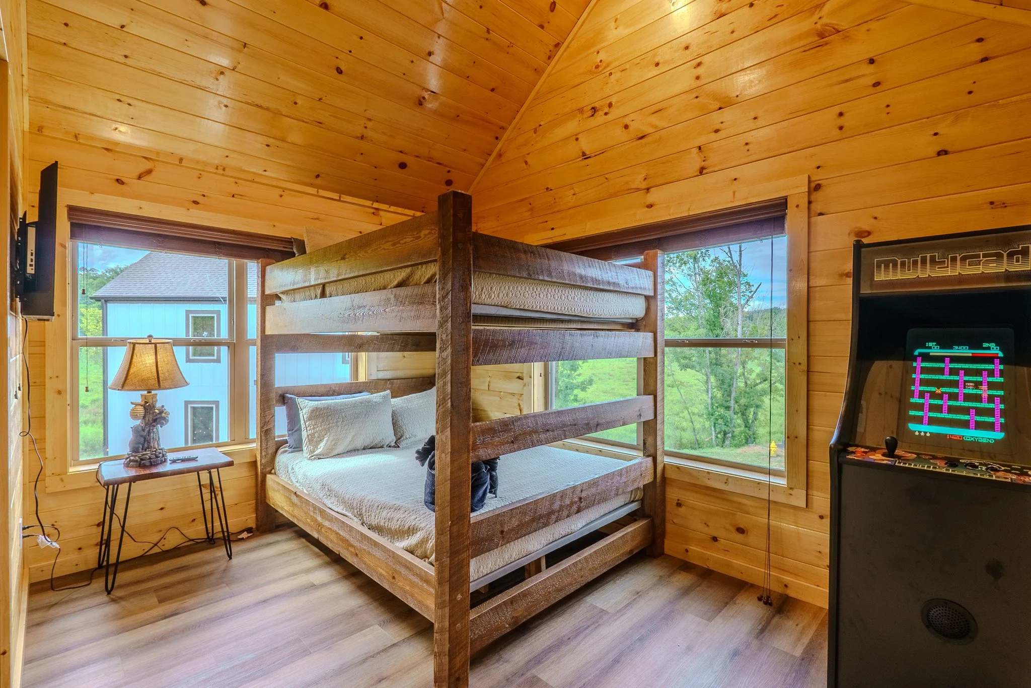 A cozy bedroom features wooden walls and ceiling, a wooden bunk bed with a twin on top and a full on the bottom, a small side table with a lamp, and a vintage arcade game machine, with two large windows showing green trees outside.
