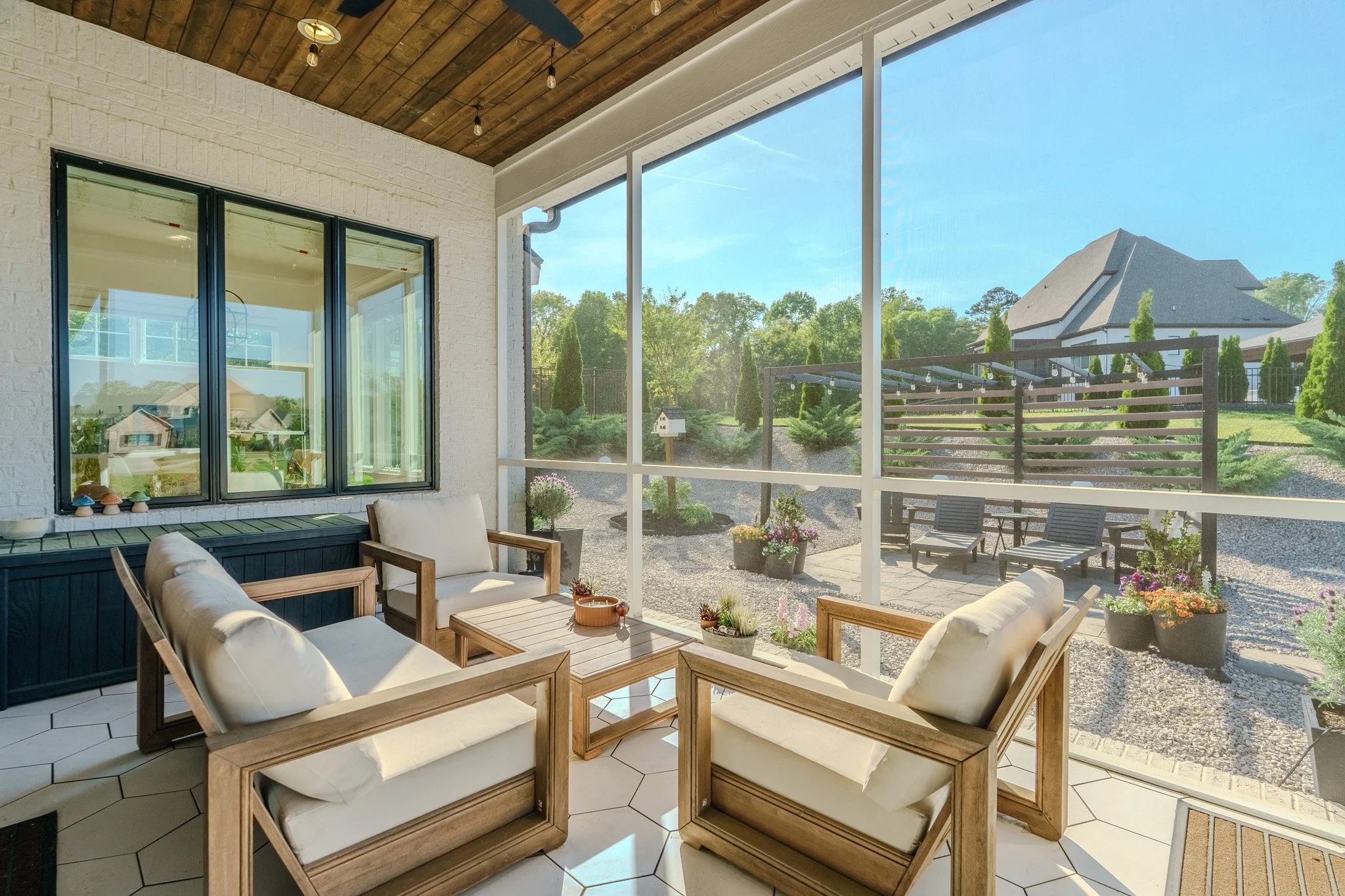 Sunlit screened porch with white brick wall, wooden ceiling, white hexagonal tiles, outdoor seating with four wooden armchairs with cushions around a wooden table, planter boxes with flowers, view of backyard with garden, patio furniture, potted plan