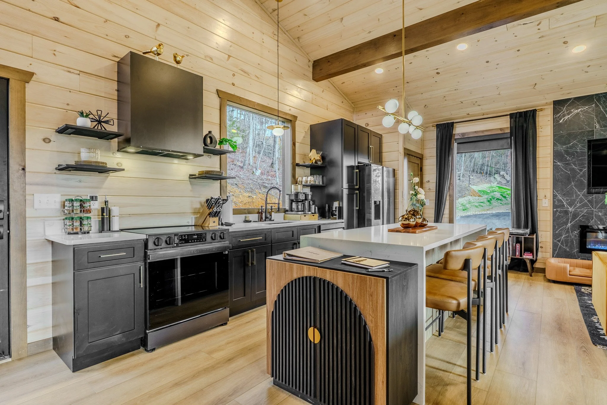 Modern kitchen with black cabinets, white countertops, wooden accents, and barstools, with large windows and a view of a wooded landscape outside.