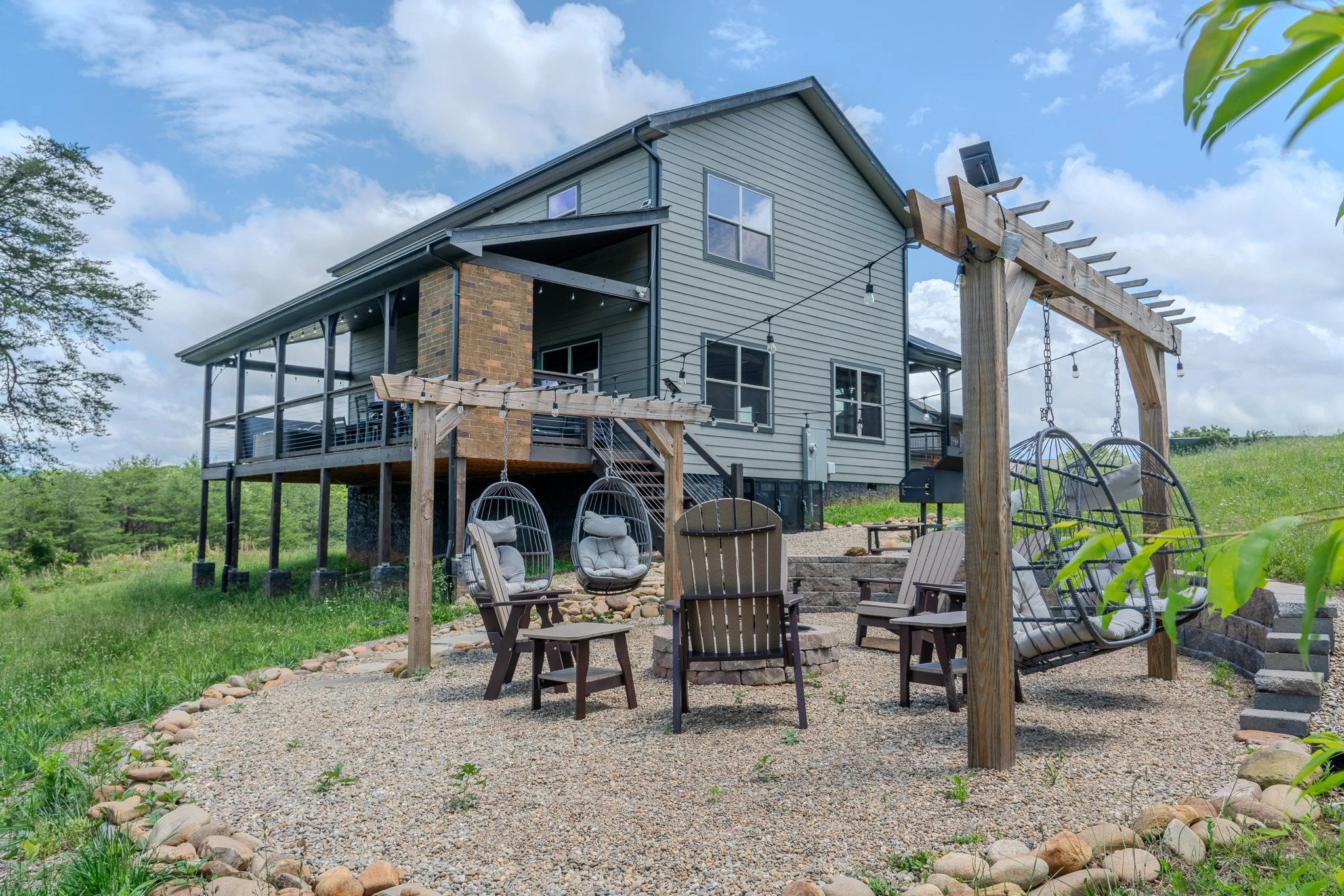 Outdoor backyard with hanging chairs, patio furniture, and a wooden swing set in front of a modern house with a deck, brick chimney, and green grass under a partly cloudy sky.