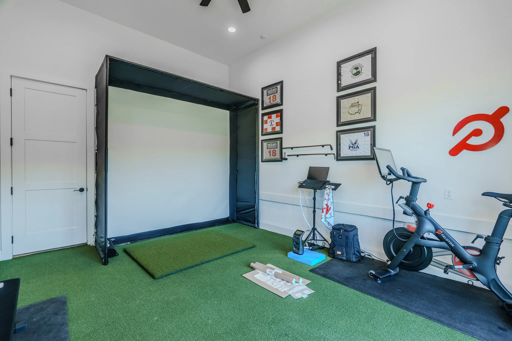 Indoor golf simulation room with a white wall, framed sports memorabilia, a golf hitting net with green turf mat, a stationary exercise bike, and various equipment and decor.